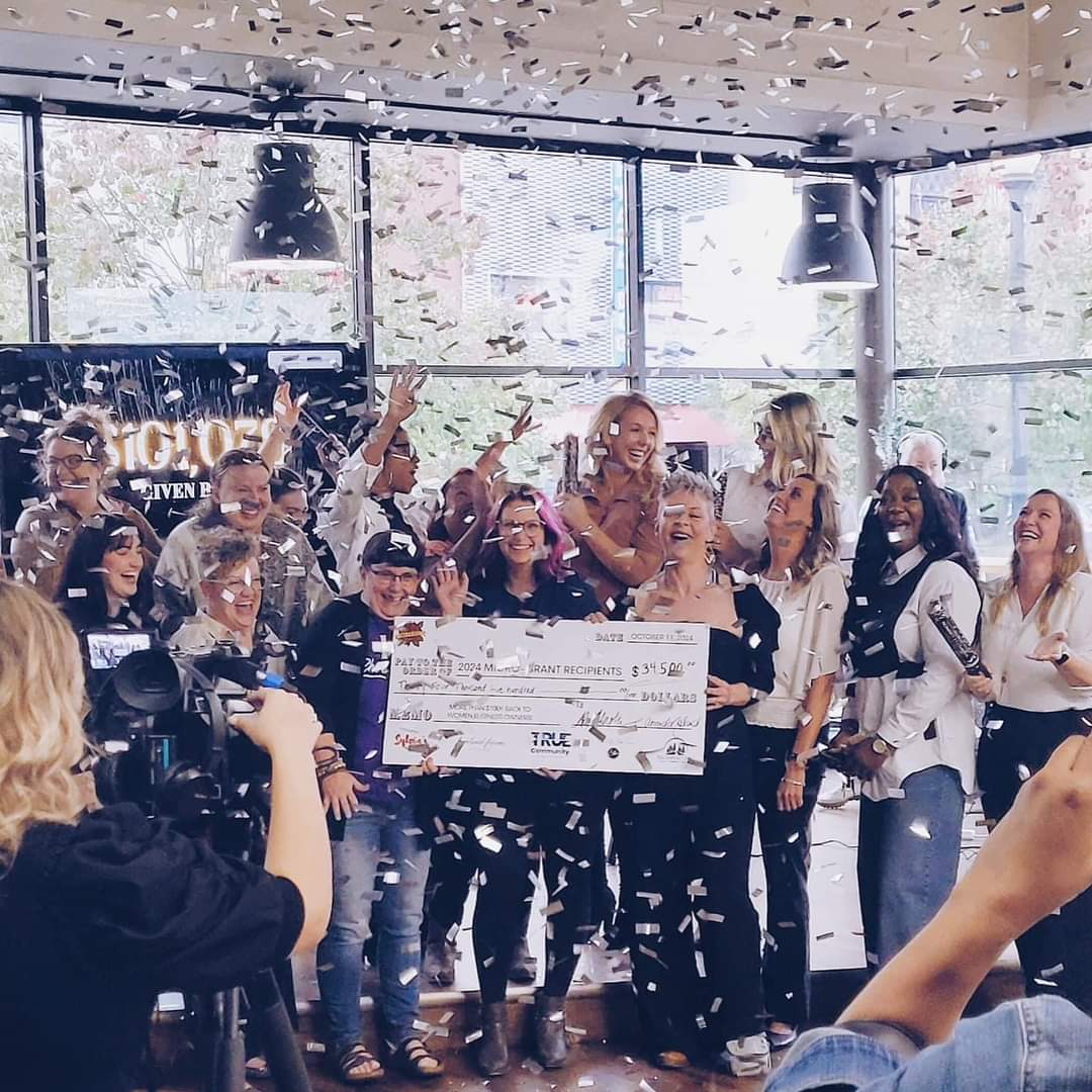 Group of people celebrating indoors with confetti, holding a large check, smiling, and raising hands, with cameras capturing the moment.