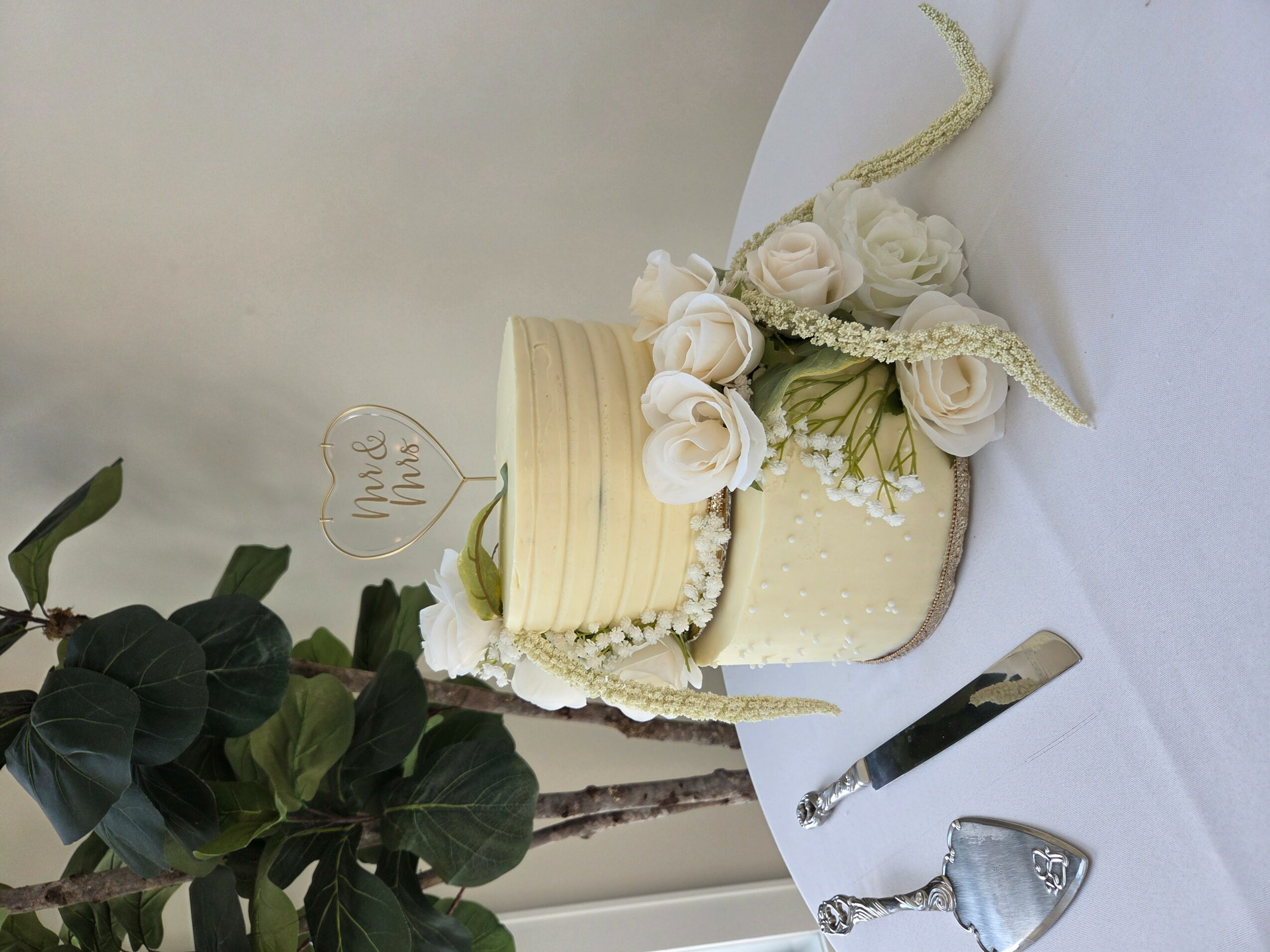 A wedding cake decorated with white roses, pearls, and a small sign, placed on a white tablecloth with a knife nearby.