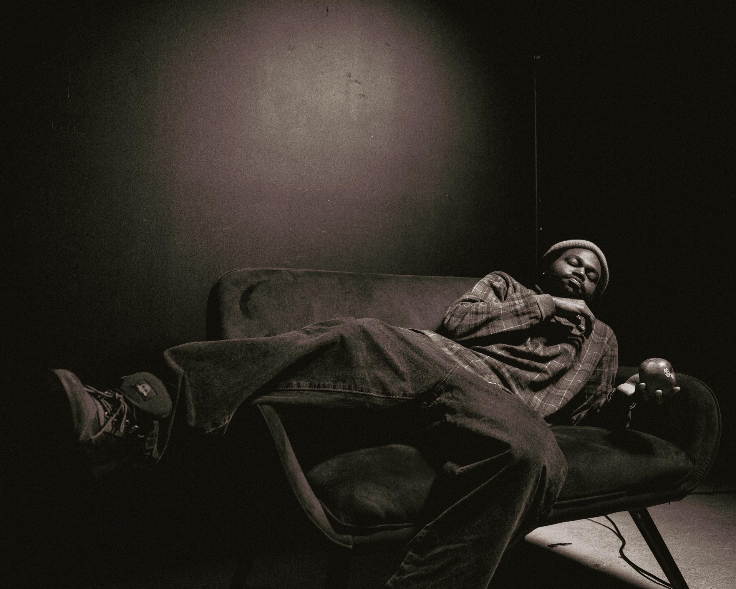 Person lying on a couch in a dark room, relaxed with arms crossed, wearing a hat and casual clothing.