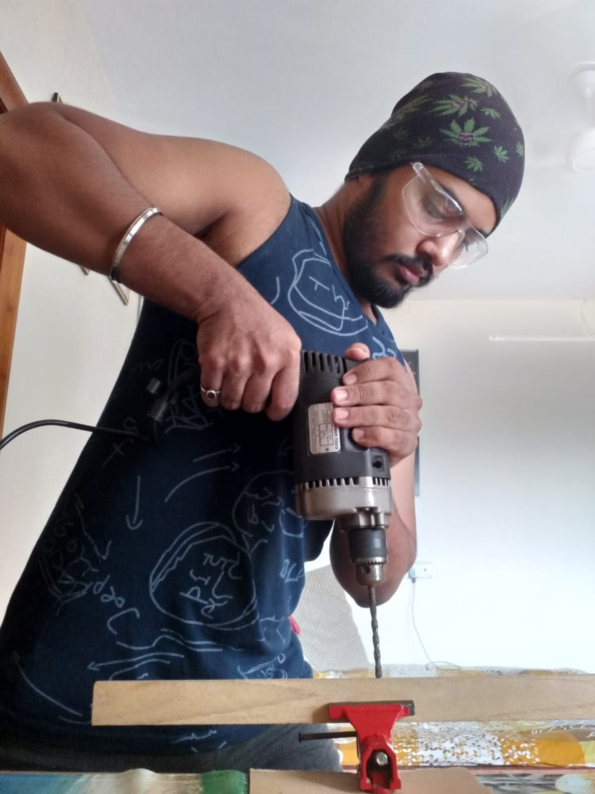 Man wearing glasses and a bandana uses a drill on a wooden surface, indoors with a white ceiling and wall.