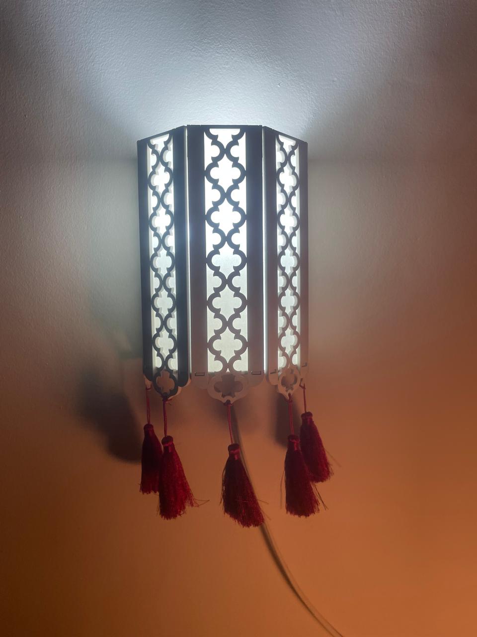 Decorative wall lamp with black and white geometric pattern, red tassels hanging from bottom, mounted on a wall, light shining upward.