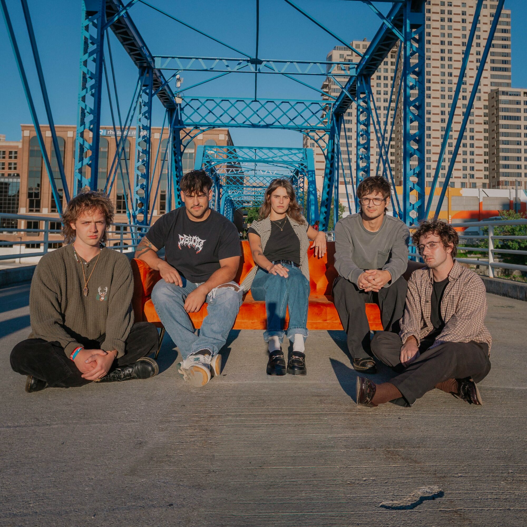 Five young people sitting and standing on a bridge with blue metal framework and city buildings in the background.
