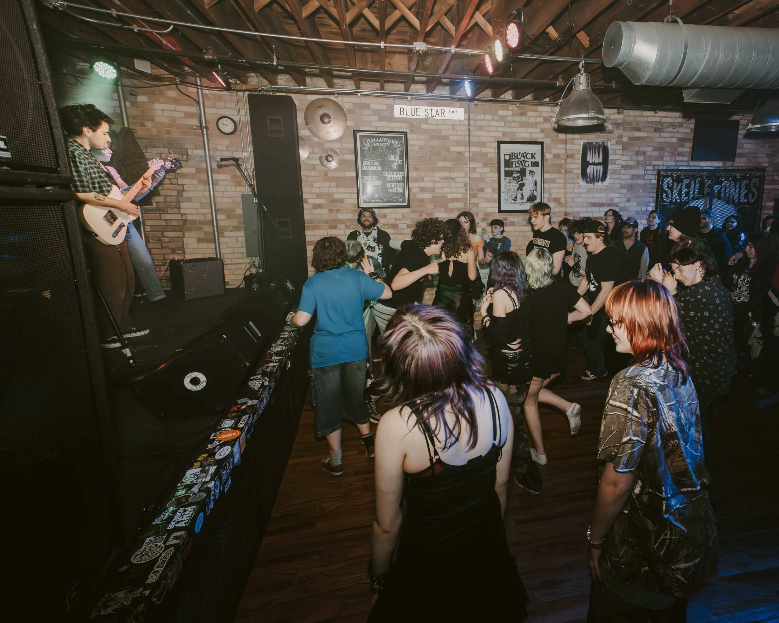 People dancing and watching a musician perform on stage in a brick-walled venue with posters and a sign reading 'Skull & Bones'.