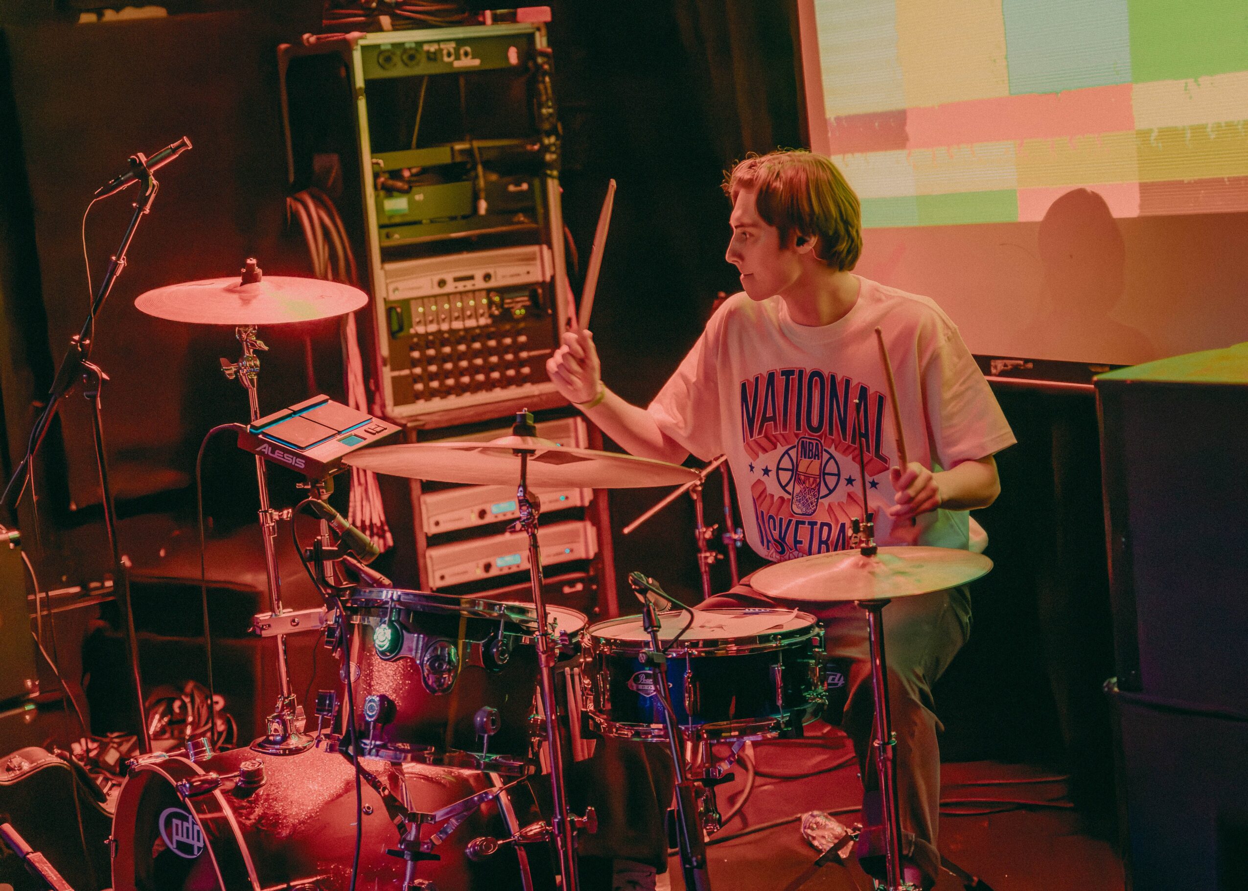Person playing drums on stage with electronic equipment and a colorful screen in background.