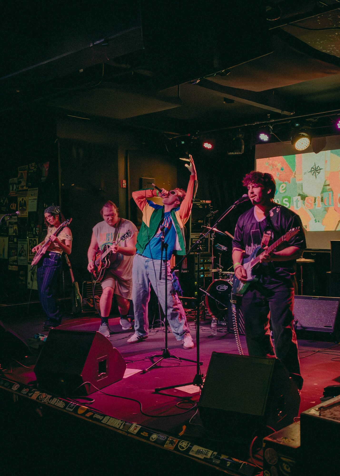 Band performing on stage with four members, colorful background, and musical equipment, in a dimly lit venue.