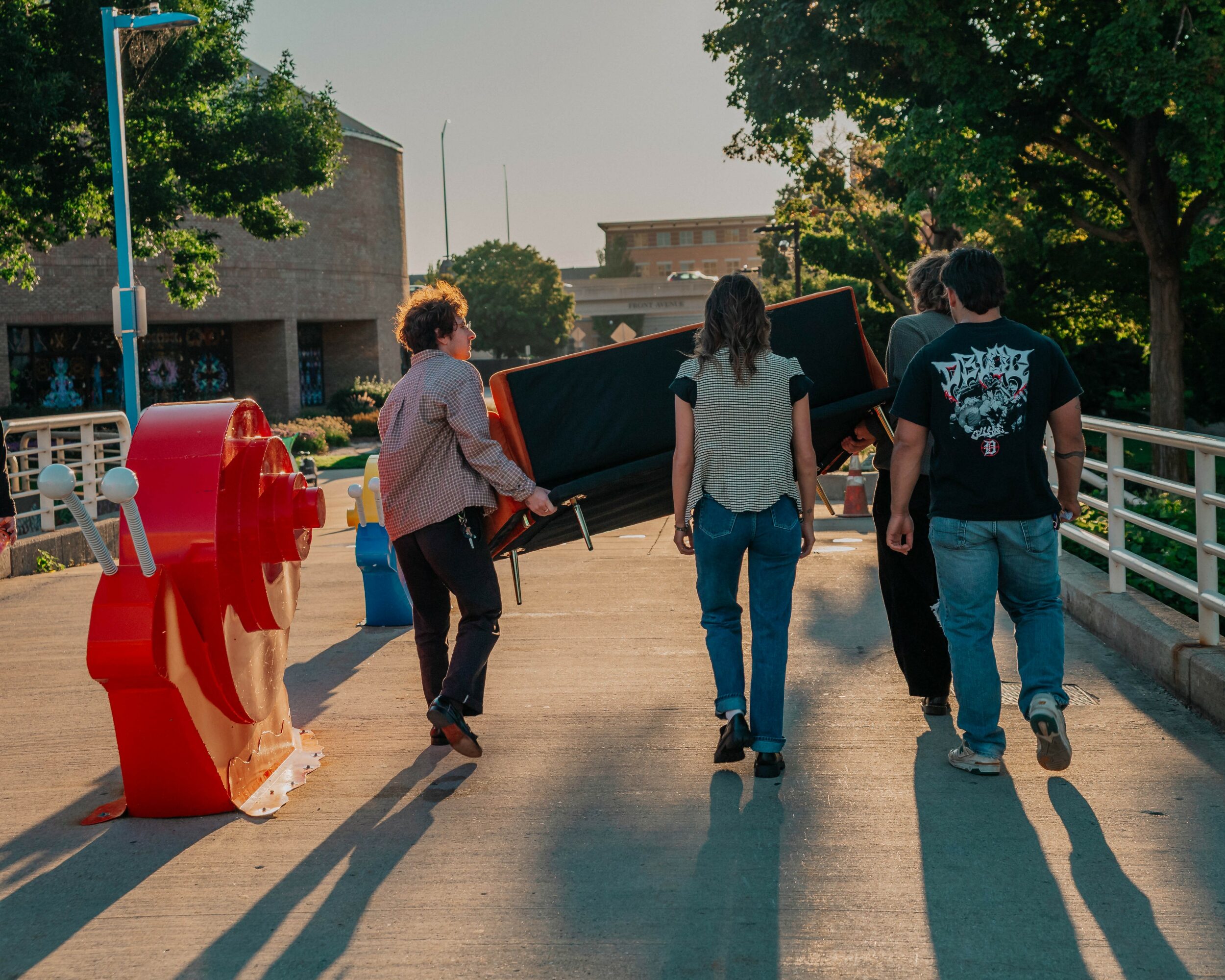 Four people carry a large object on a bridge with trees and buildings in the background.