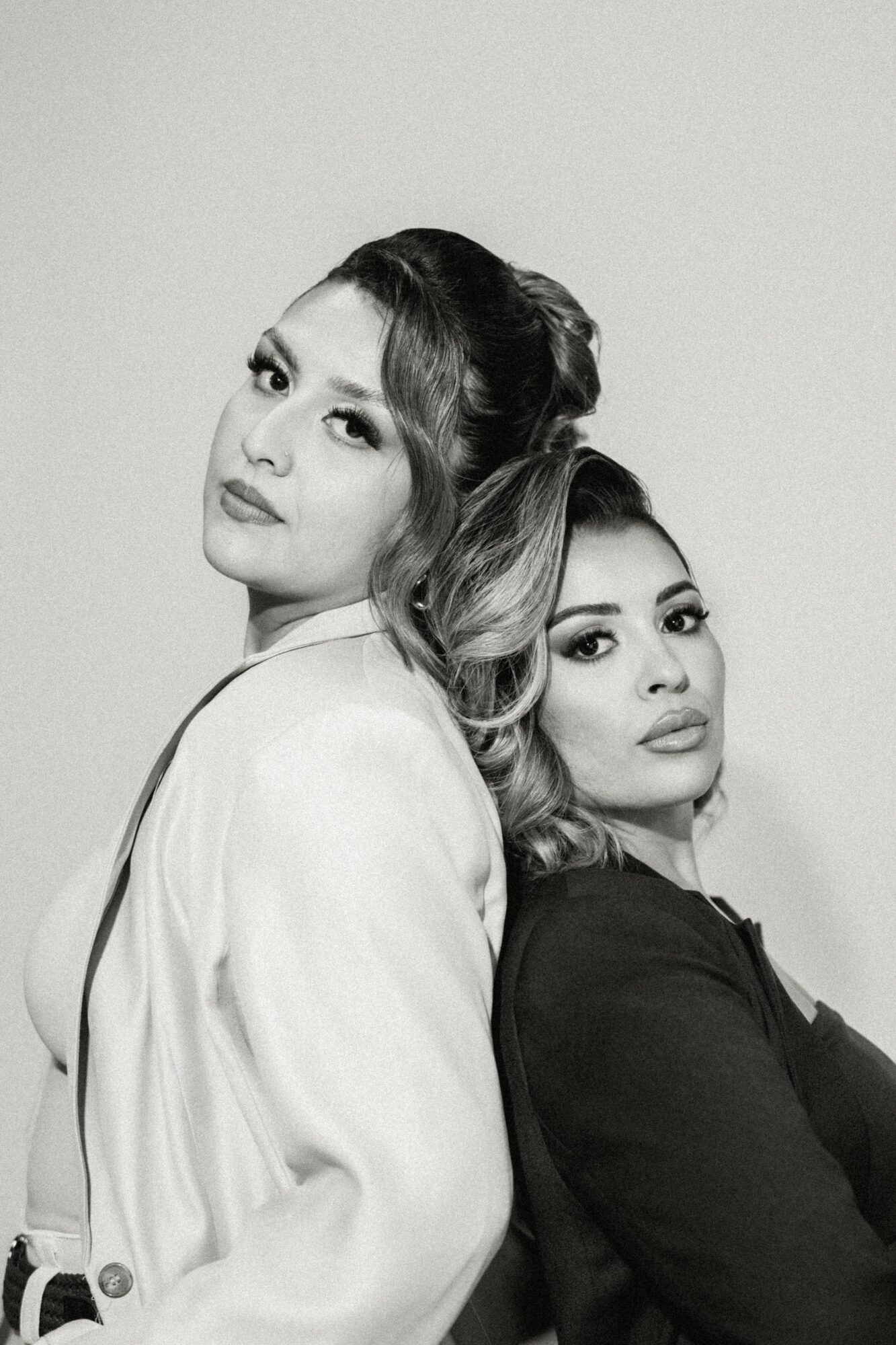 Two women pose closely, one in a white blazer and the other in a dark top, against a plain background.