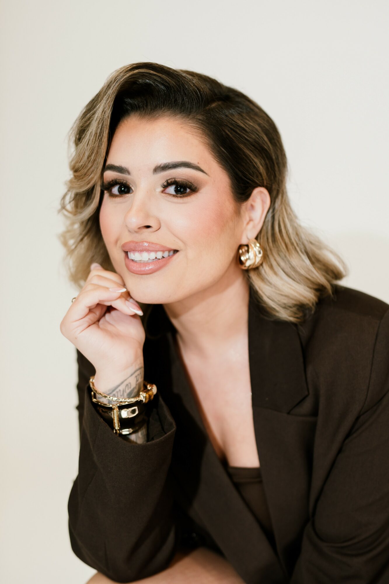 Woman with short wavy hair, wearing a black blazer and gold jewelry, smiling and resting her chin on her hand.
