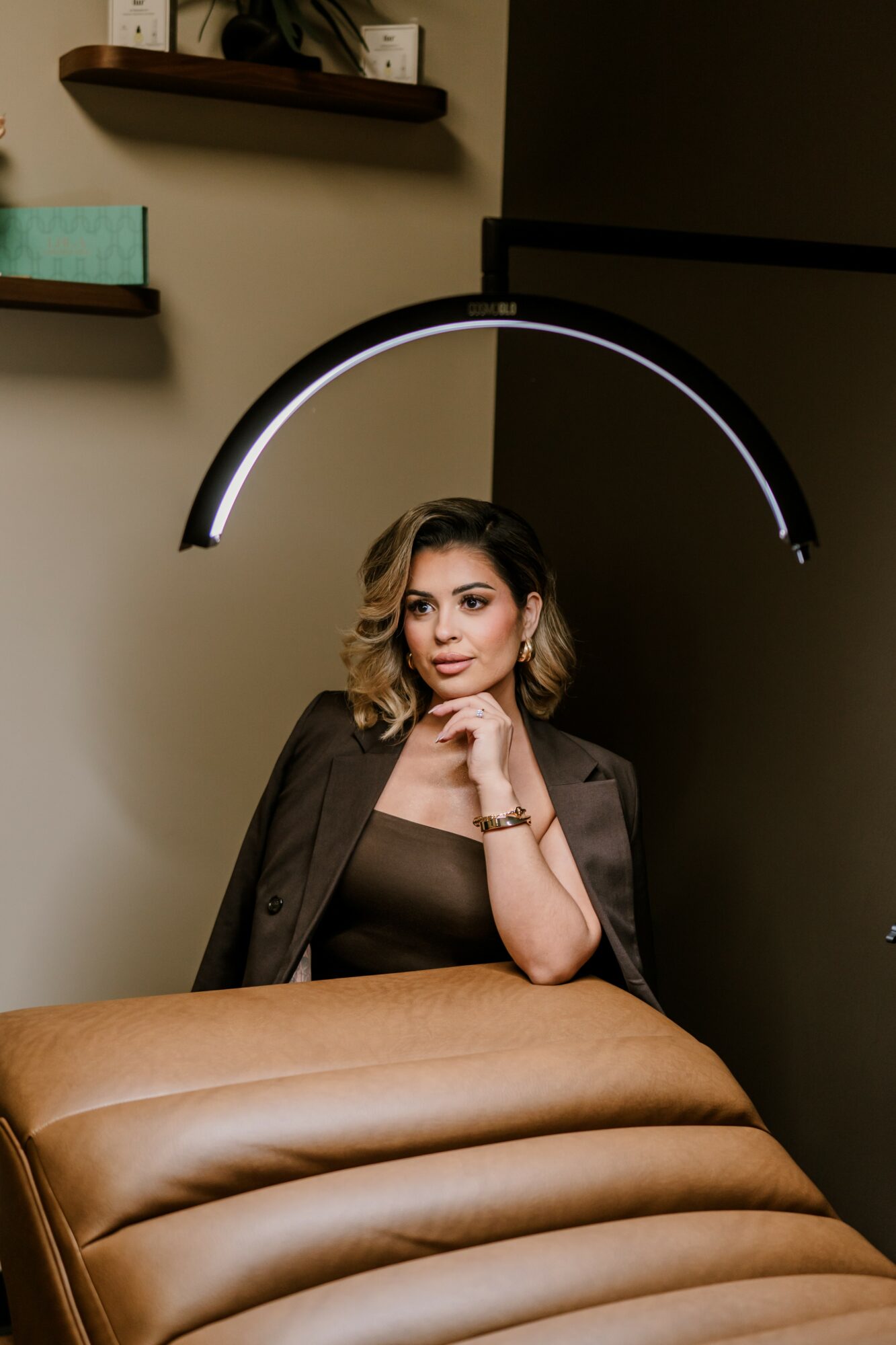 Woman with shoulder-length hair sitting at a table under a modern hanging light fixture, with shelves on the wall behind her.