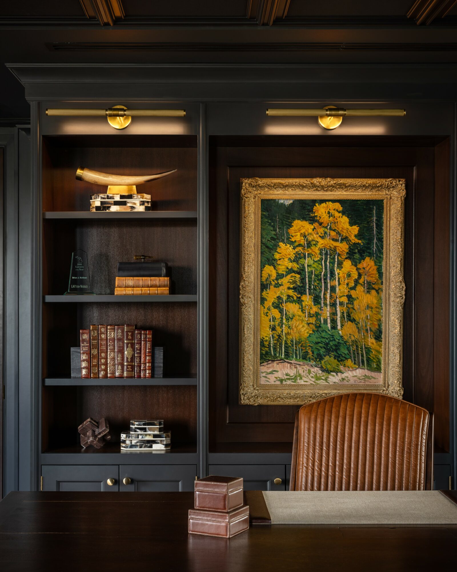 Bookshelf with a painting of yellow flowers and trees, and a brown chair in front of a desk, in a room.