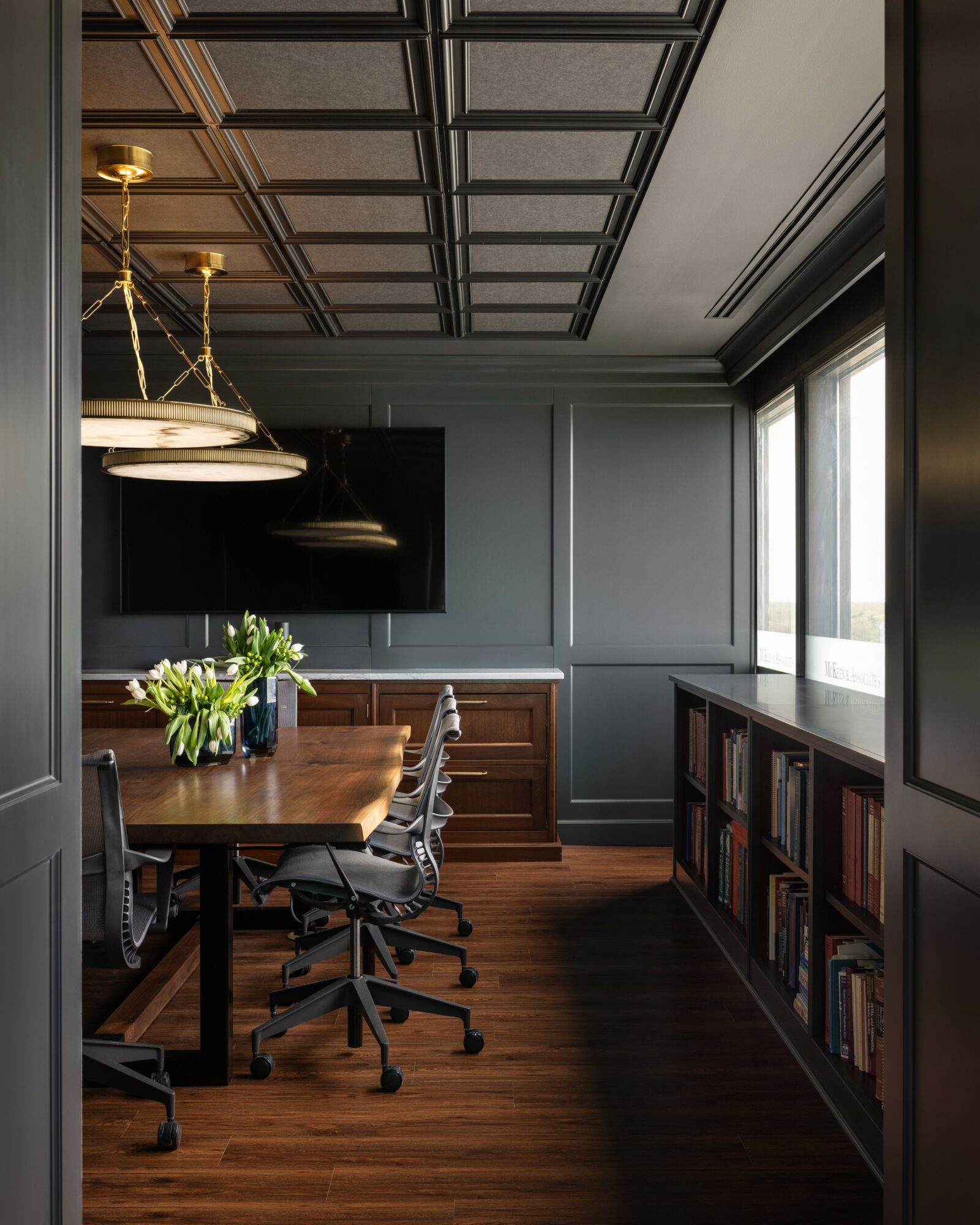 Conference room with wooden table, chairs, bookshelf, window, and ceiling lights, viewed from doorway.