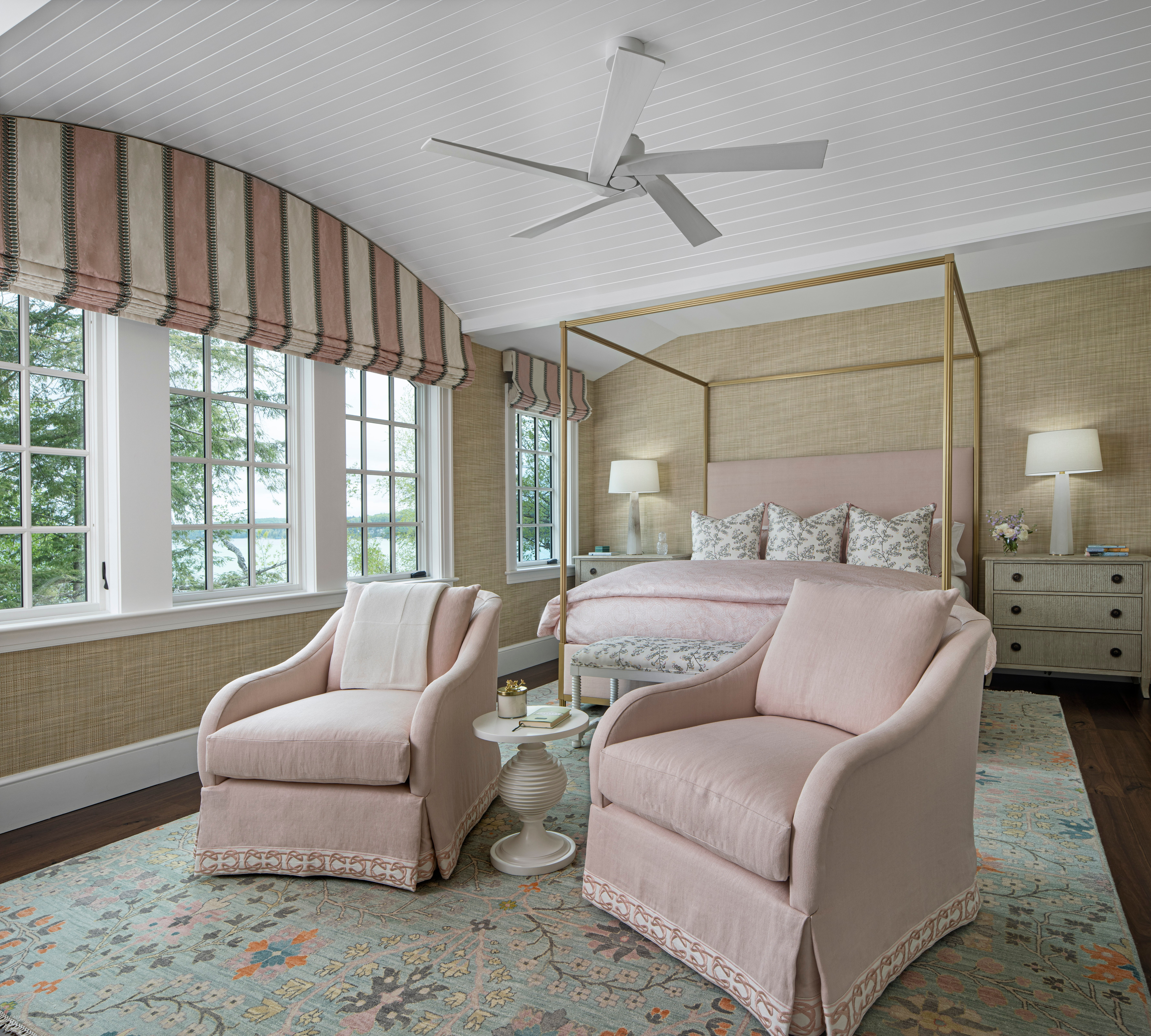 Bedroom with a canopy bed, two pink armchairs, a side table, and large windows with striped valance, ceiling fan, and lamps.