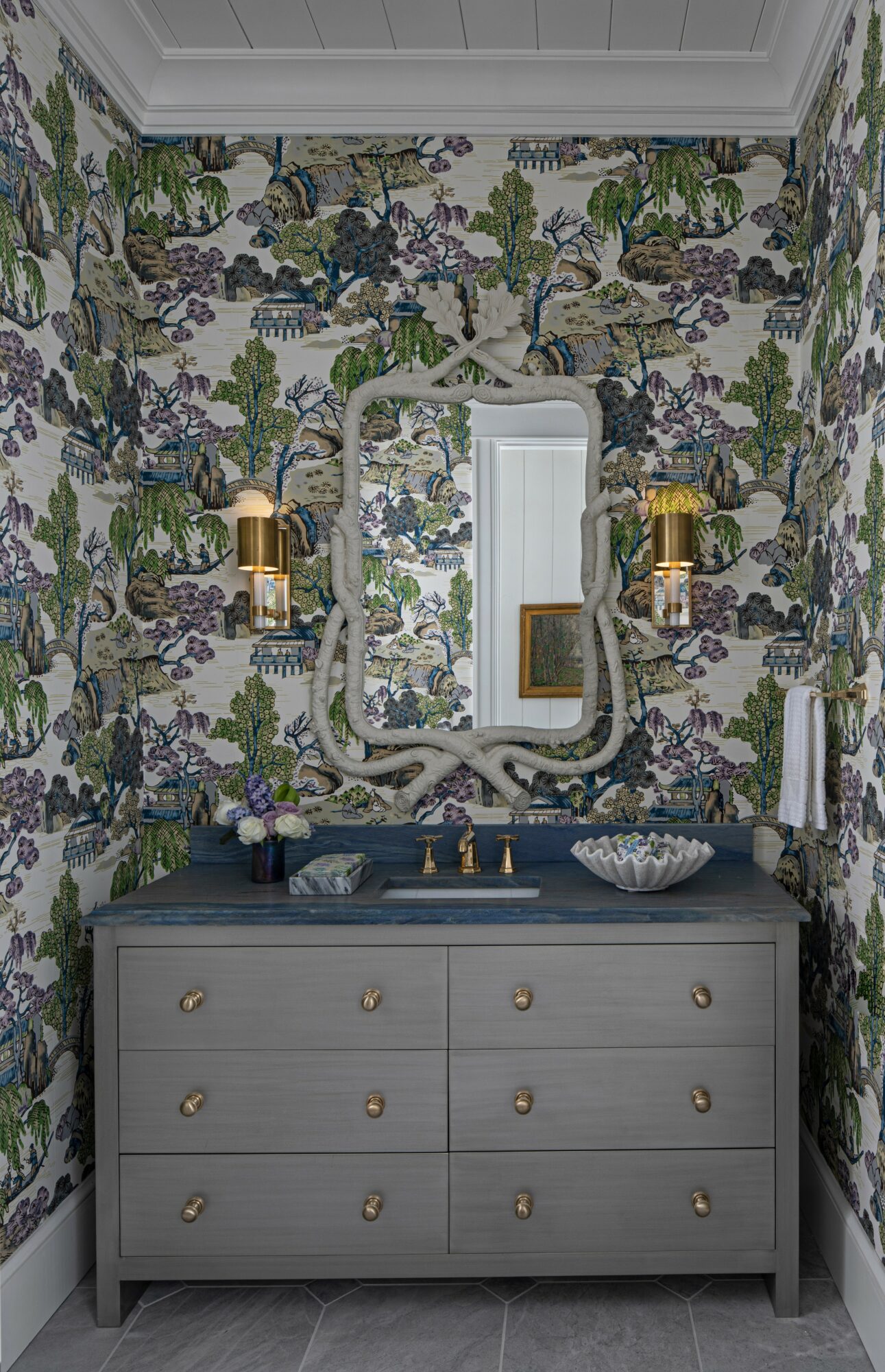 Bathroom vanity with a gray cabinet, gold fixtures, a decorative mirror, and floral wallpaper background.