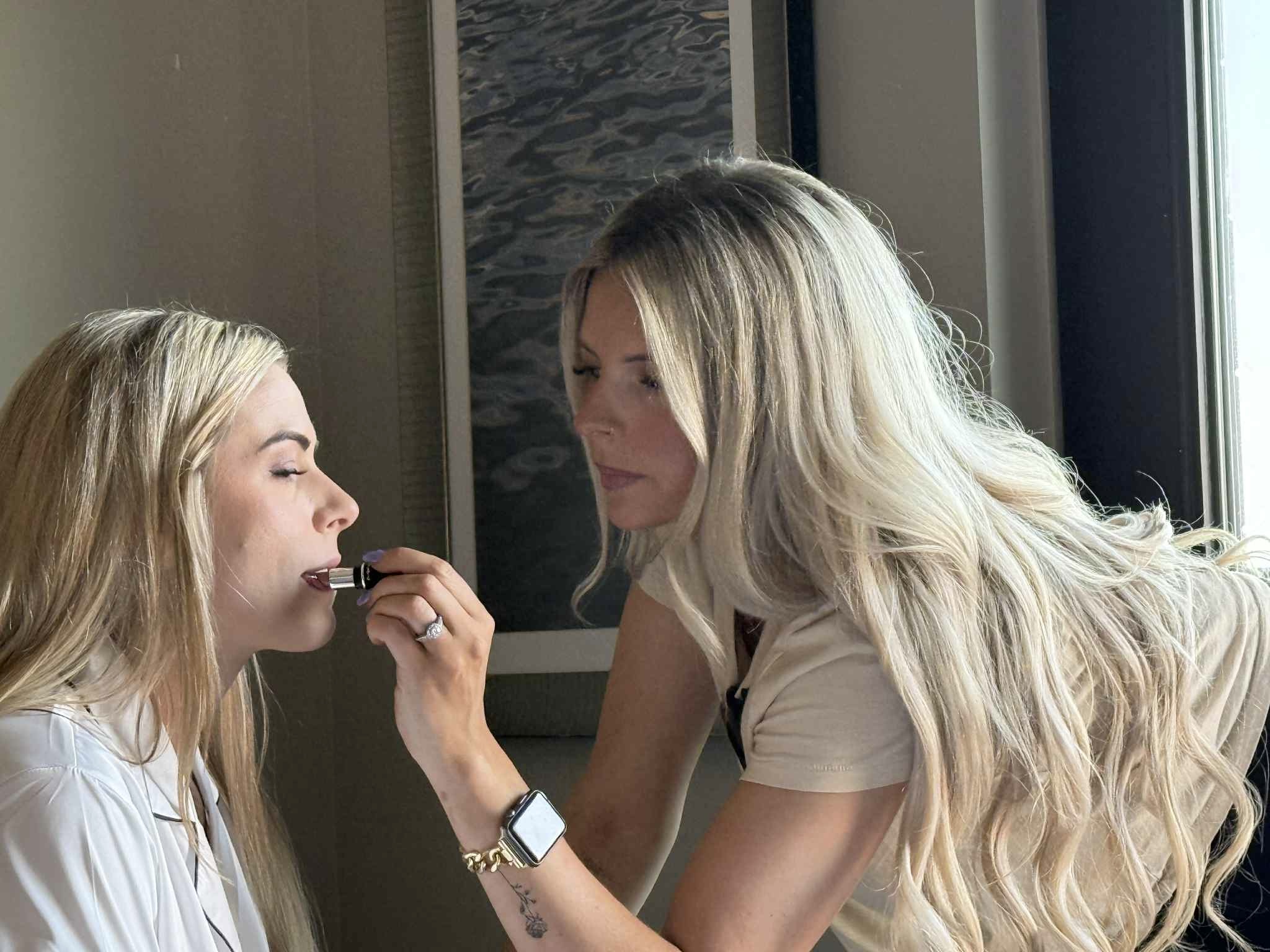 Two women with long blonde hair, one applying makeup to the other, indoors near a window with a view outside.