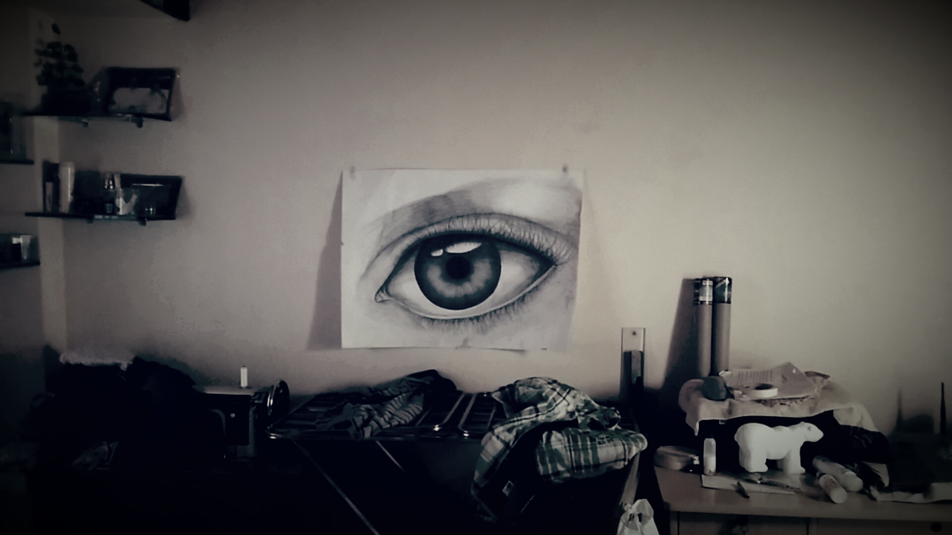 Close-up of a detailed eye drawing on a wall, with cluttered objects below, in a room.