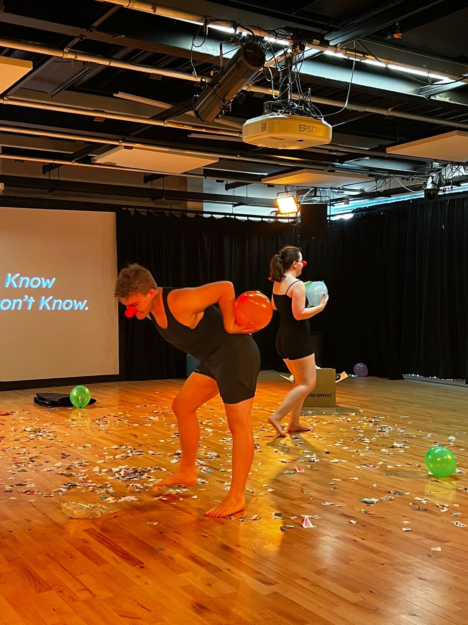 Two people on stage with a black curtain, one holding a ball, the other with a ball on their back, wooden floor, scattered objects.
