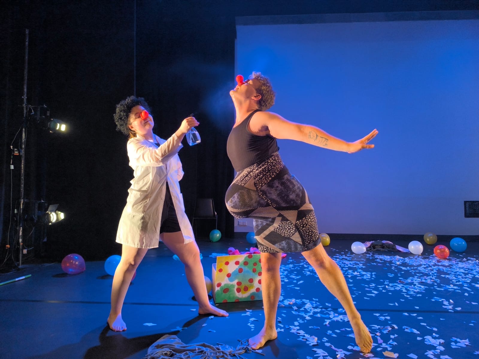 Two performers on stage, one in a white coat and the other in a black top and patterned skirt, engaging in a dance or act.