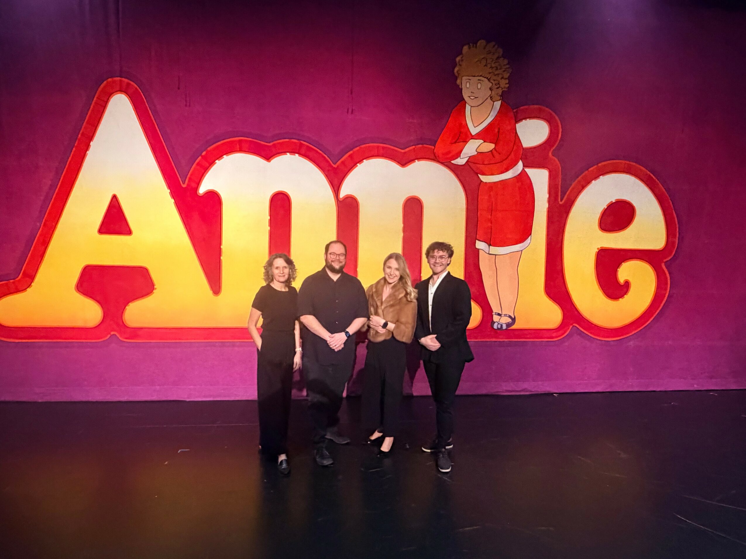 Four people standing in front of a large colorful sign with the word 'Annie' and a cartoon girl on a pink background.