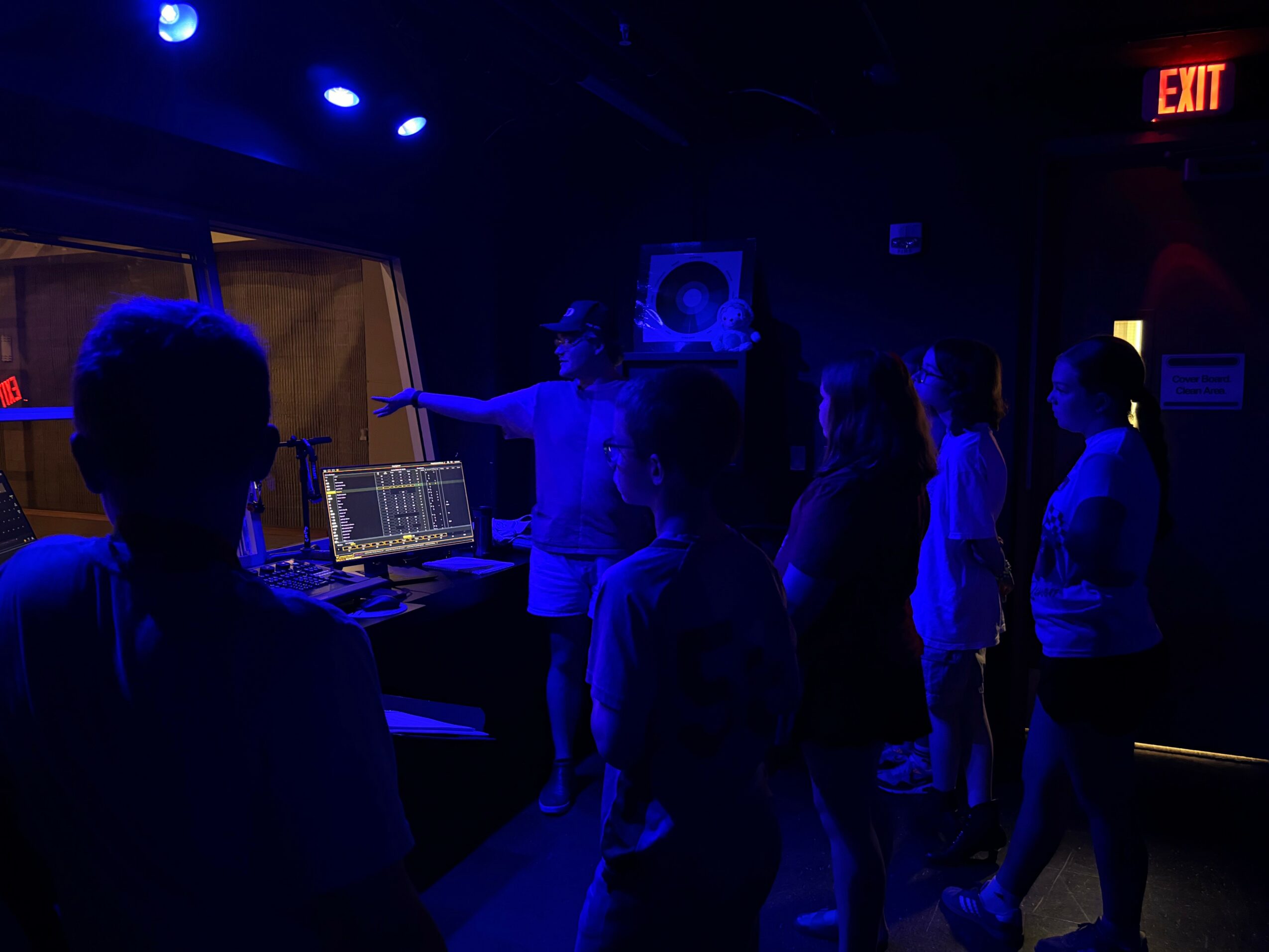 Group of people in a dark room with blue lighting, some facing a person pointing, with equipment on a table nearby.