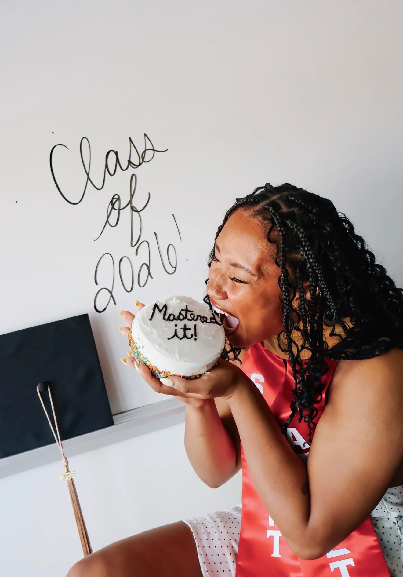 Young girl with curly hair holding a cake with a message, smiling, in a classroom with writing on the wall.