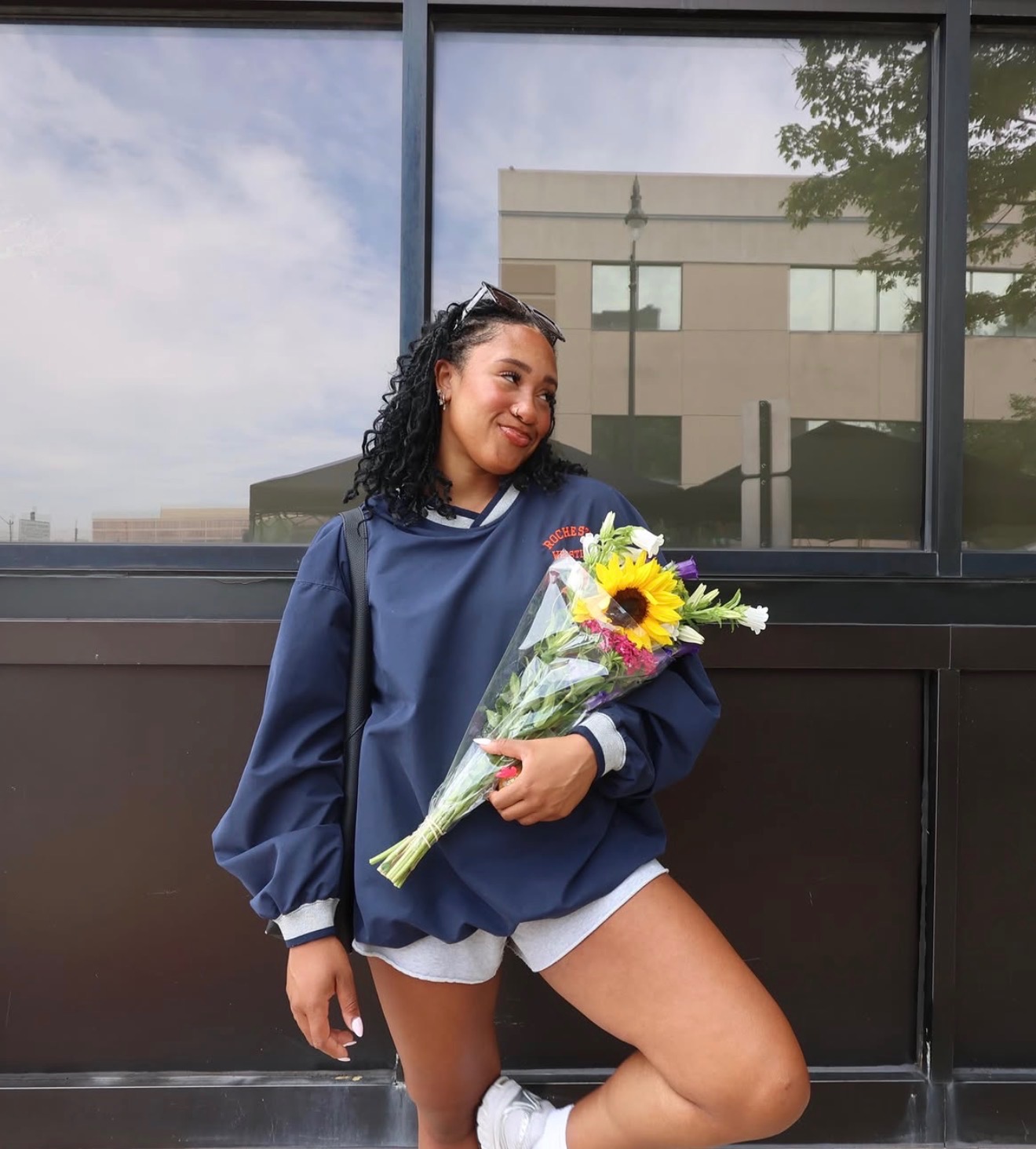 Young woman with curly hair holding a bouquet of flowers, standing outdoors in front of a building with large windows.