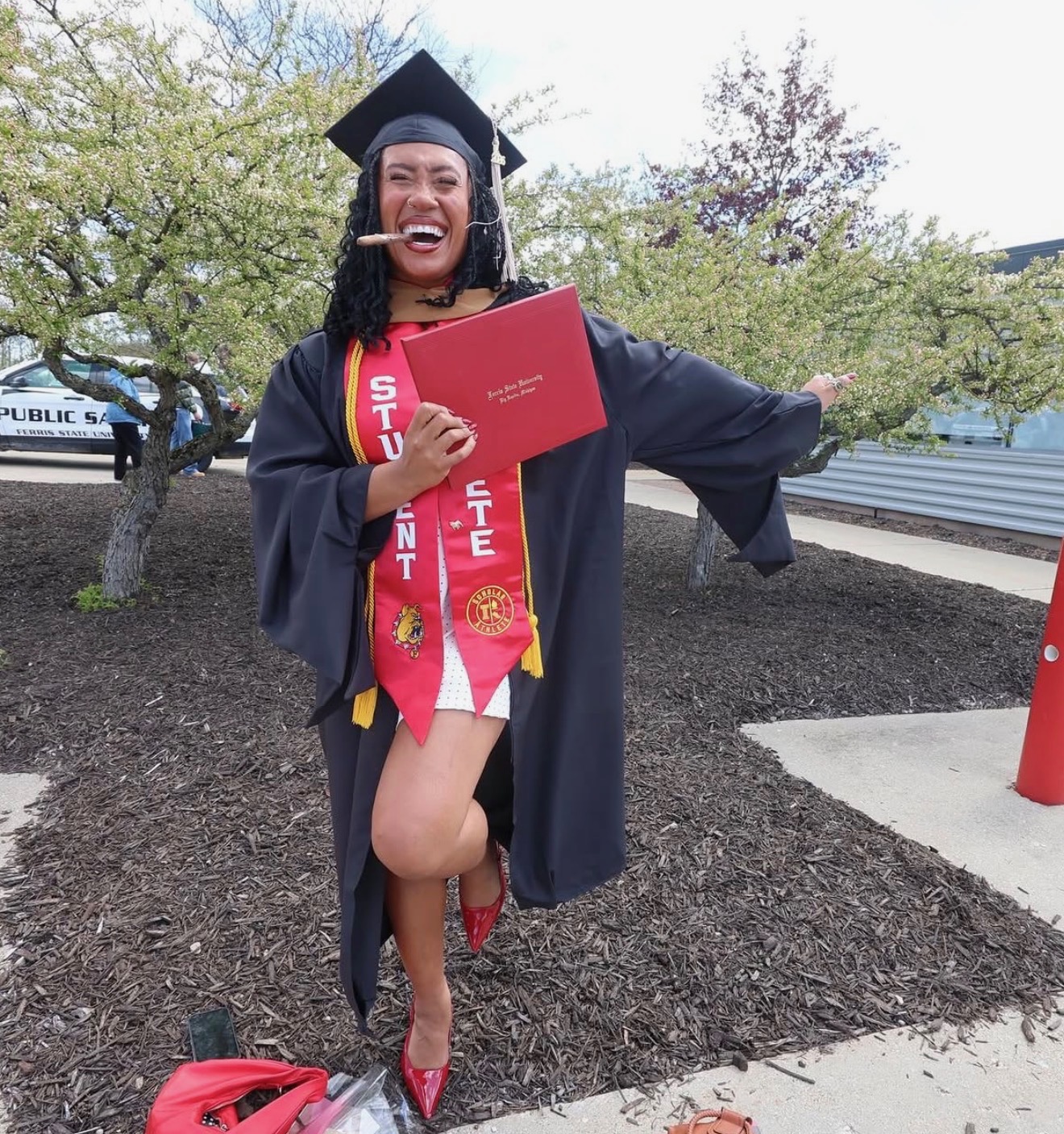 Graduate in cap and gown holding diploma, smiling outdoors with trees and pathway in background.