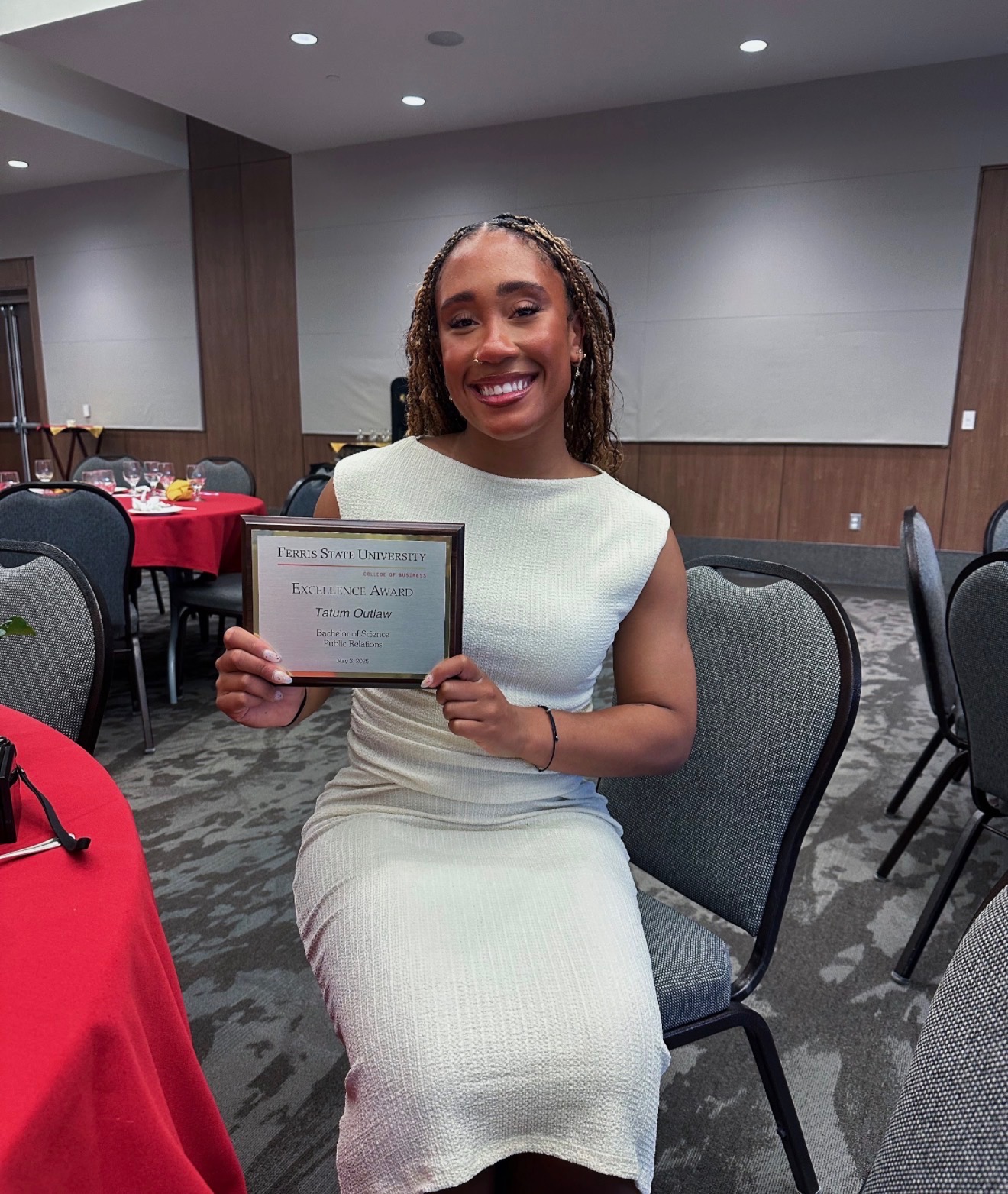 Smiling woman in white dress holding a certificate, seated at a table in a banquet room.