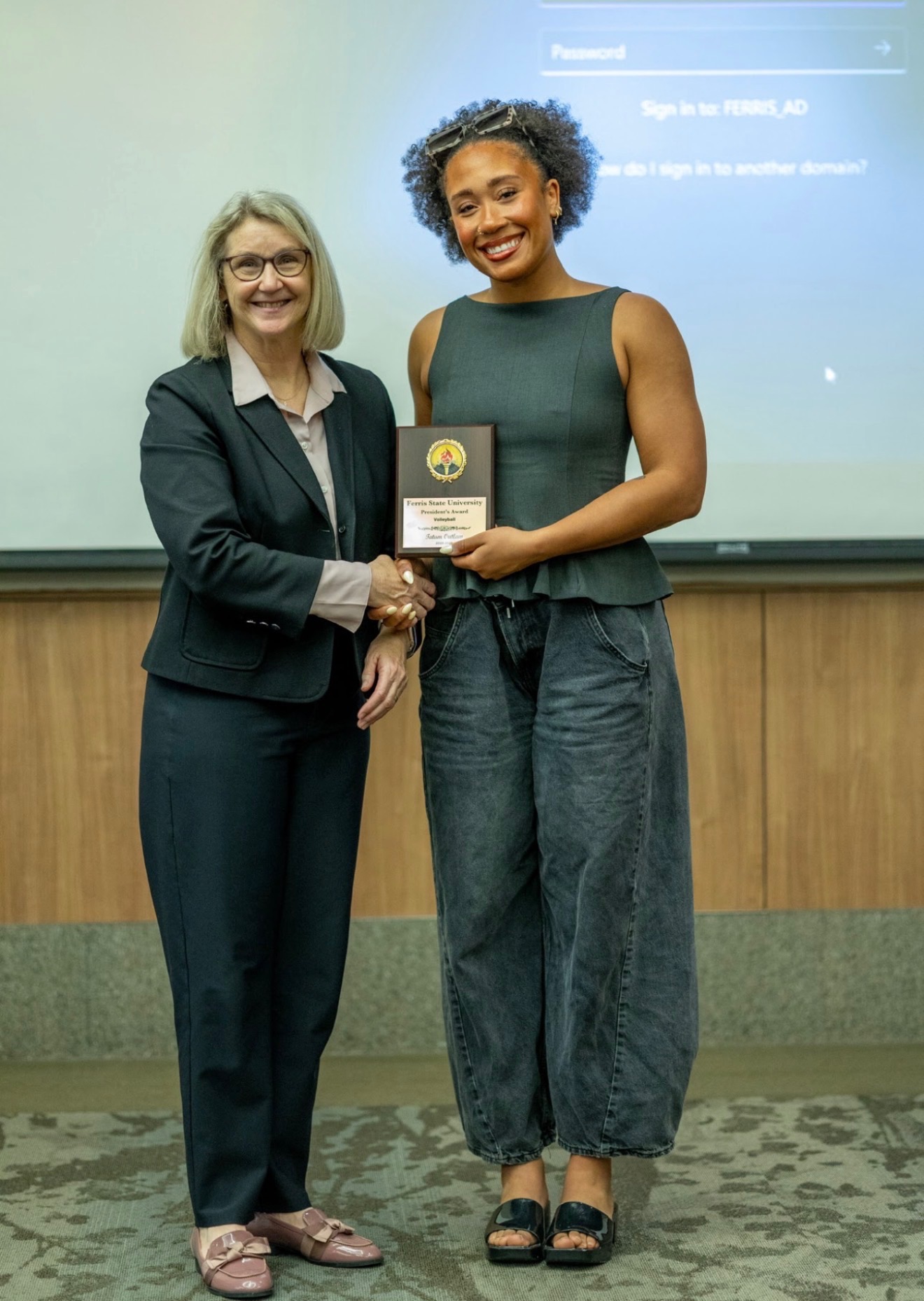 Two women standing in front of a presentation screen, one holding a plaque, smiling, in a professional setting.