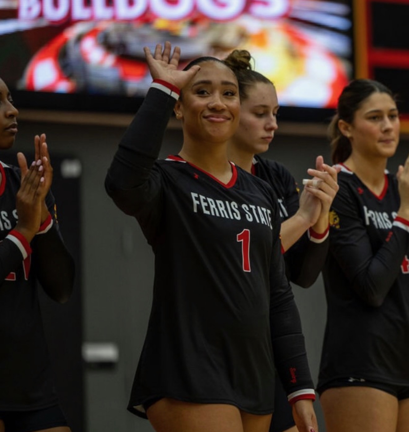 Group of female volleyball players in black jerseys, one smiling and waving, in an indoor sports arena.