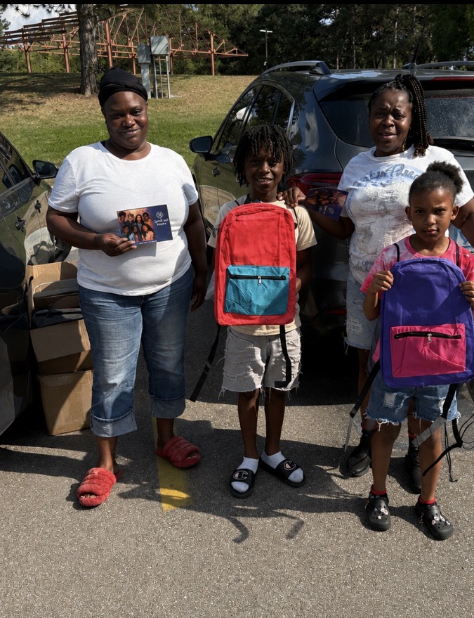 Four people standing outdoors in front of cars, holding backpacks and a photo, with trees and a fence in background.