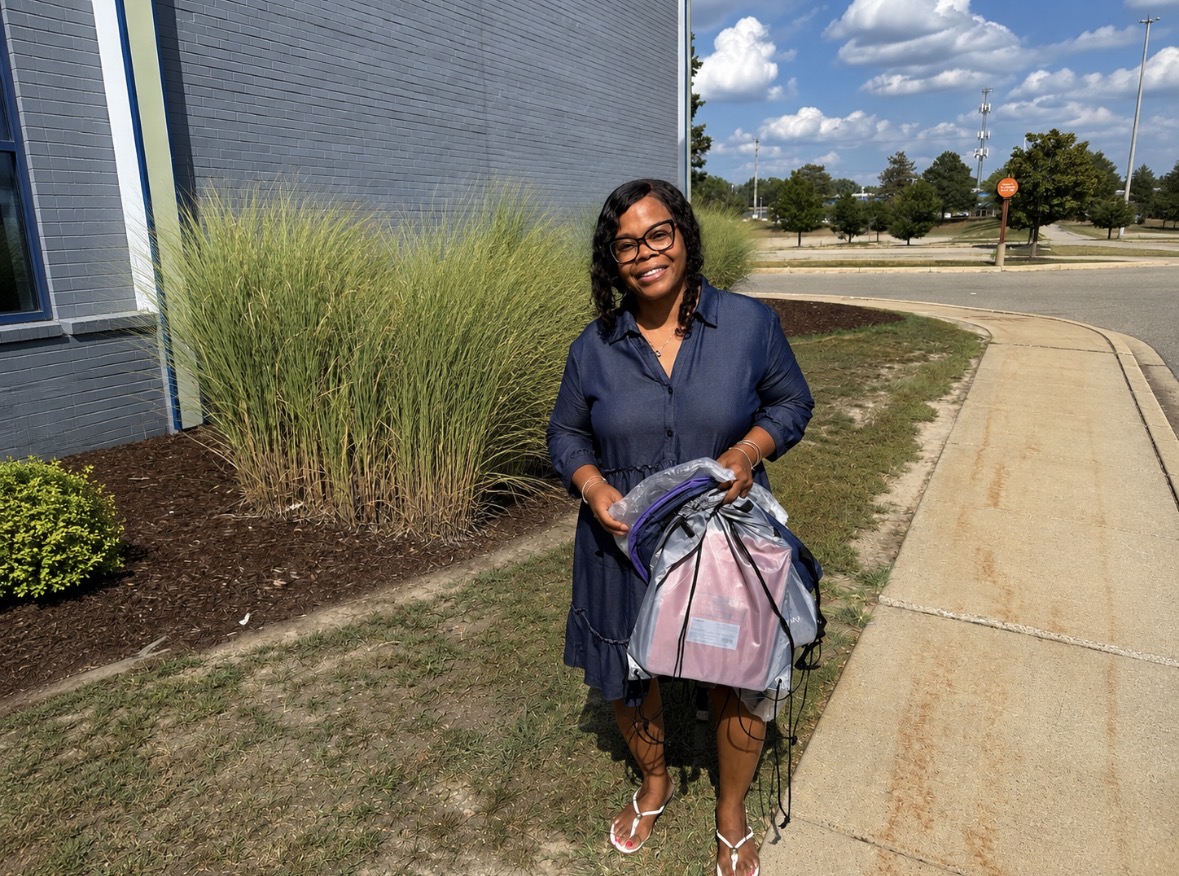 Woman standing outdoors near a building with grass and trees, holding a backpack, smiling, sunny day.