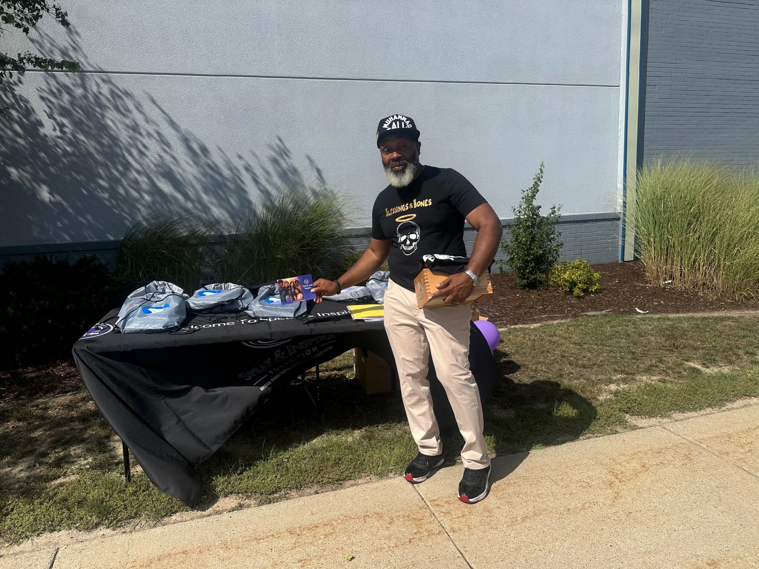 Man standing next to a table with items outdoors, near a building and grass, holding a small object in his right hand.