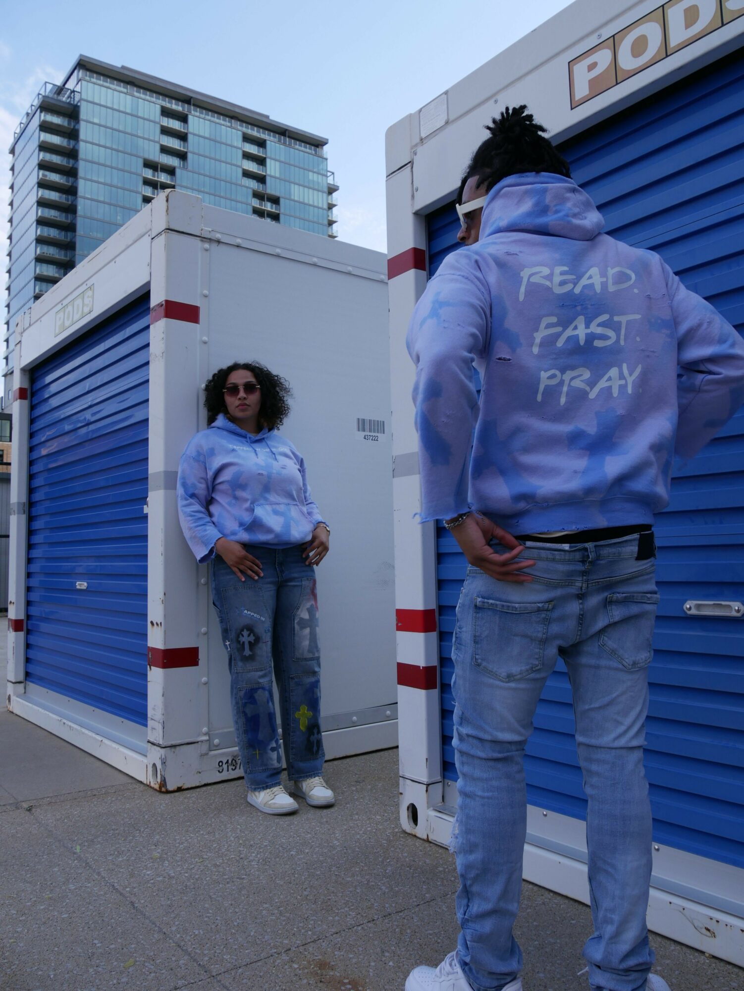 Two people standing outdoors near a large white and blue container, with a tall building in the background.