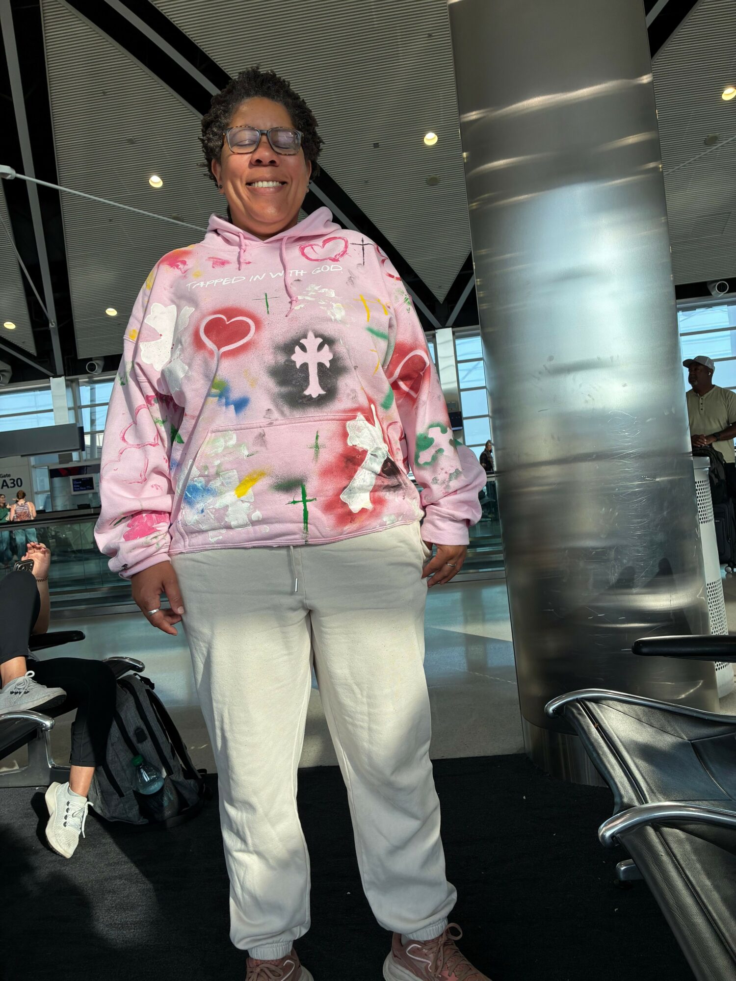 Person standing indoors, wearing a pink hoodie with various designs, white pants, and white shoes, with a backpack on the floor.