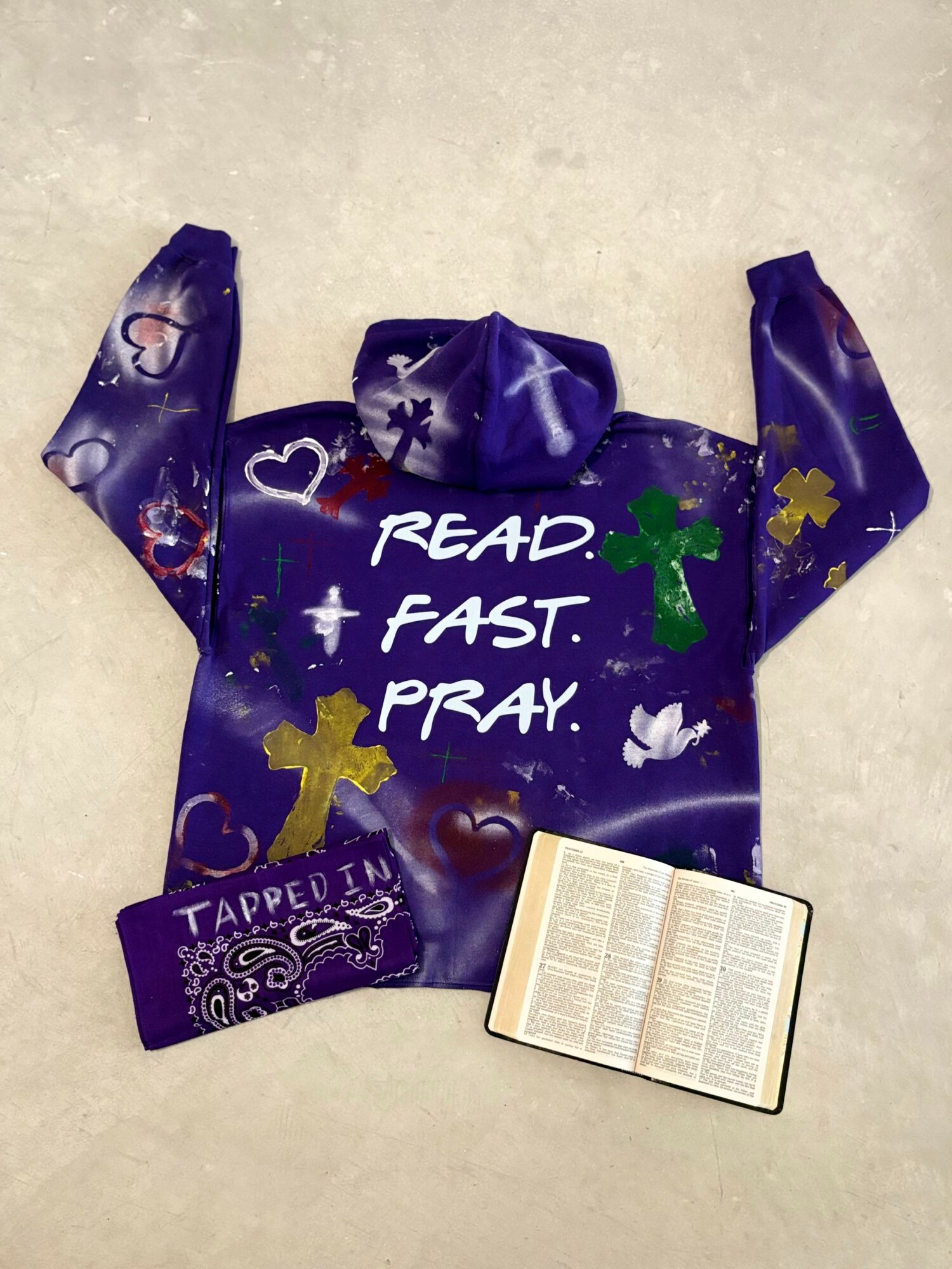 A purple sweatshirt with colorful designs and text, a Bible, and a purple cap on a light surface.