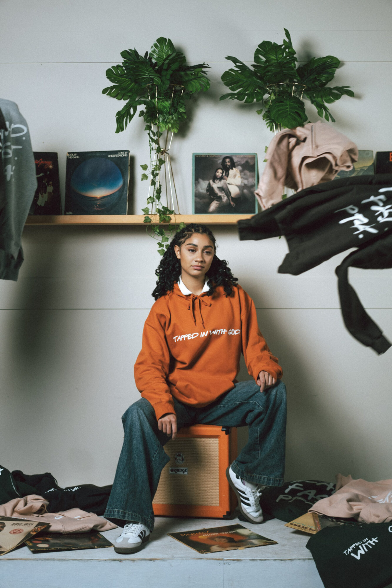 Young girl with curly hair in an orange hoodie and jeans sitting on an orange box in a room with plants and framed photos behind her.