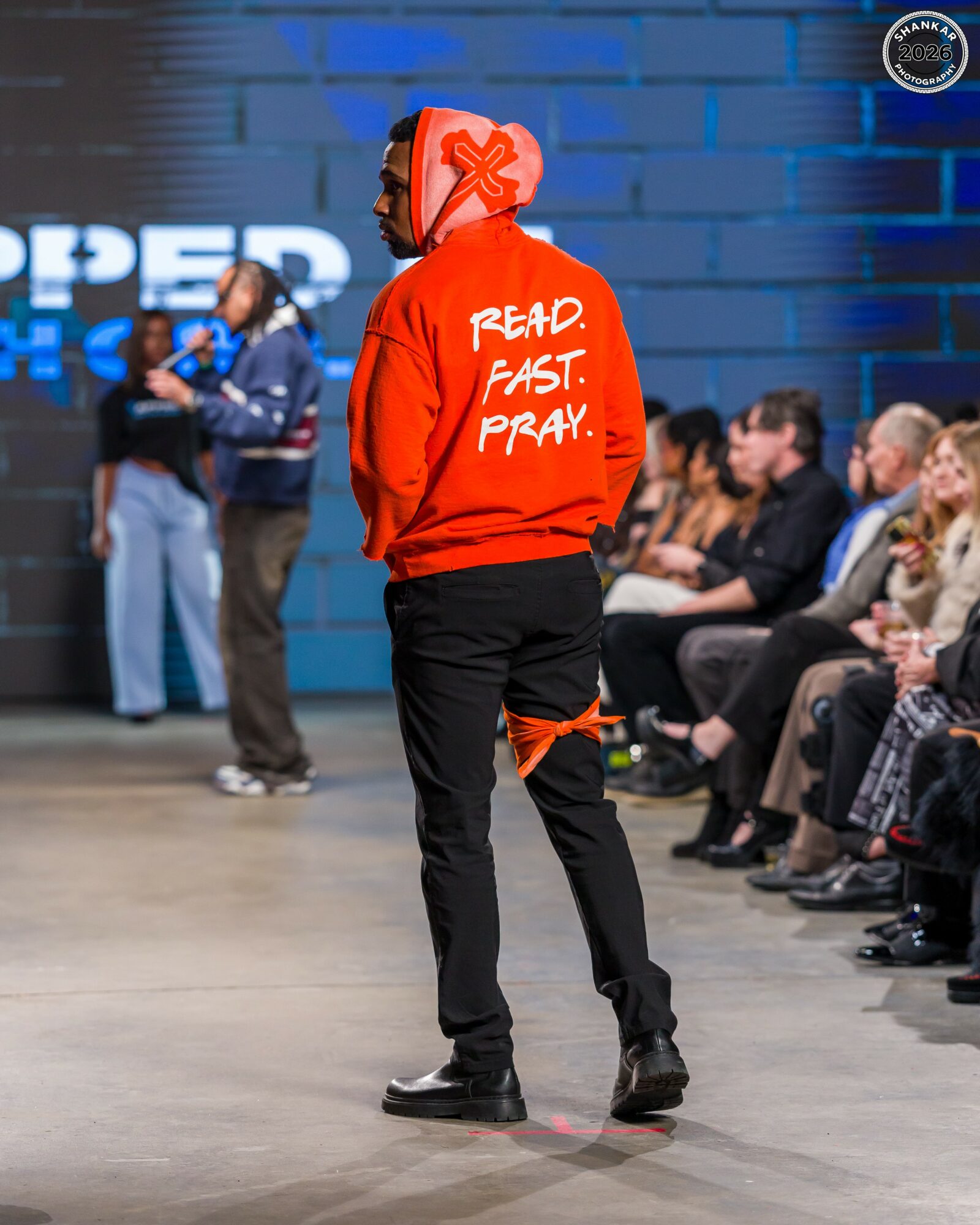Person standing with back to camera, wearing red hoodie with white text, in an indoor event with seated audience.