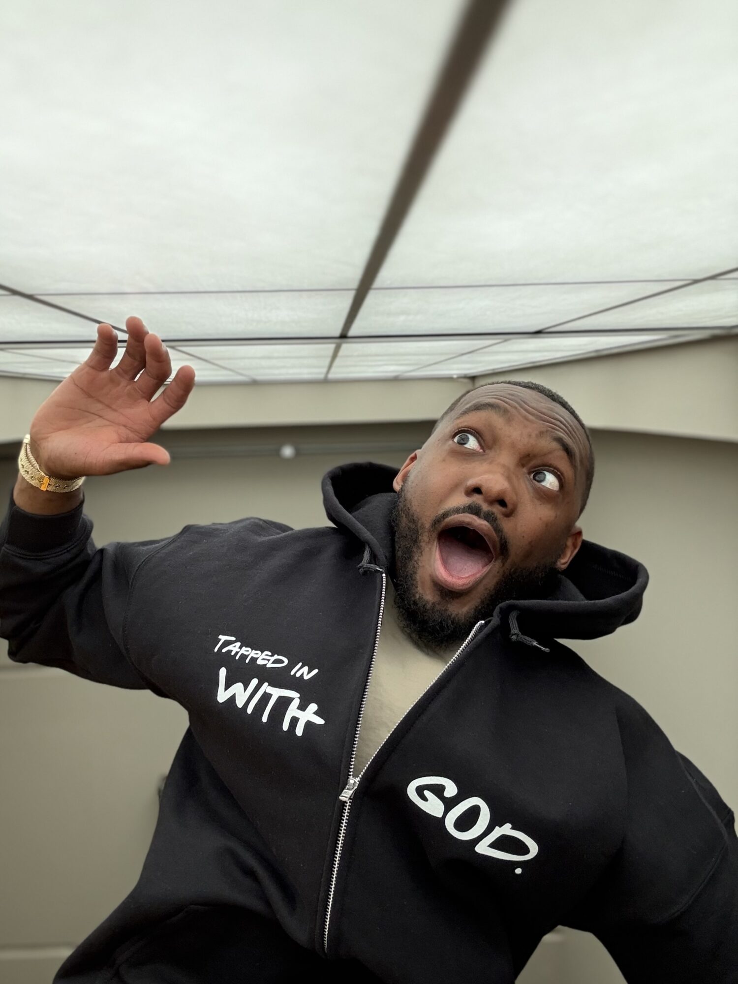 Man with surprised expression, wearing a black hoodie, raising his hand, in an indoor setting.