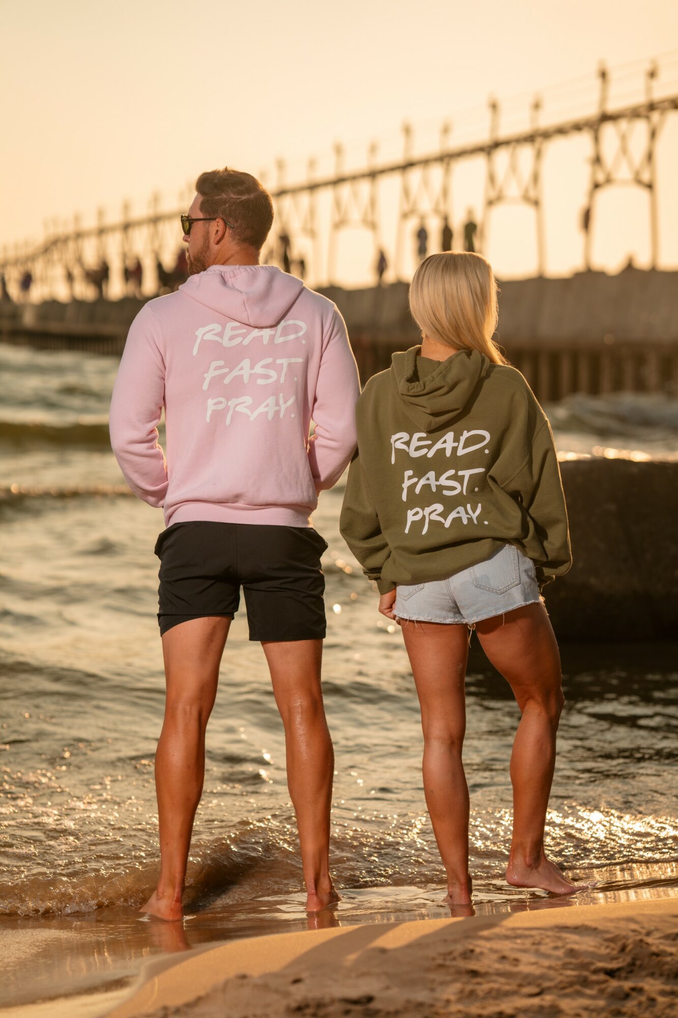 Two people stand on a beach near water, wearing hoodies with text, with a bridge in the background at sunset.