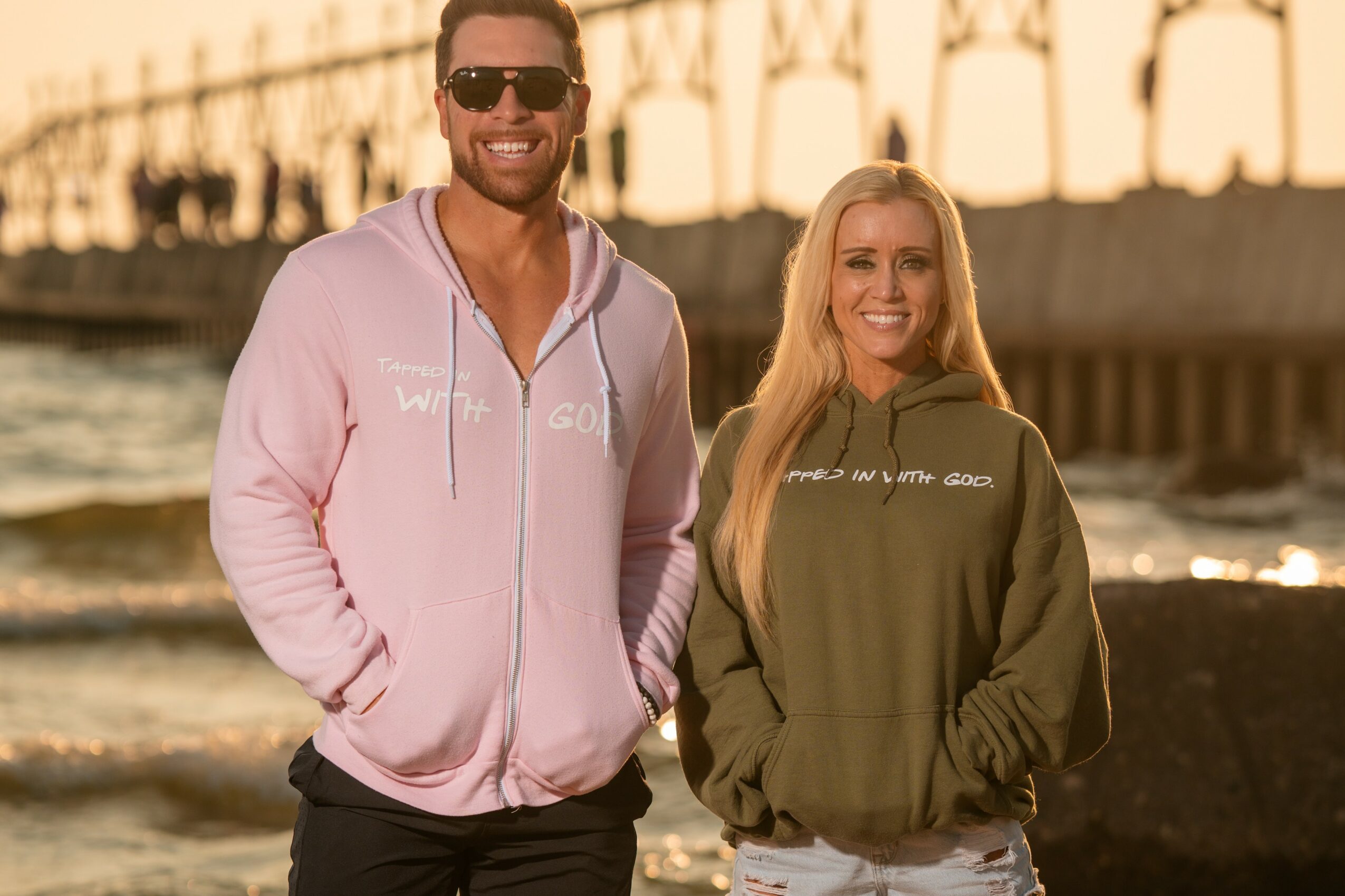 A man and woman stand on a beach with a pier in the background, smiling at the camera during sunset.