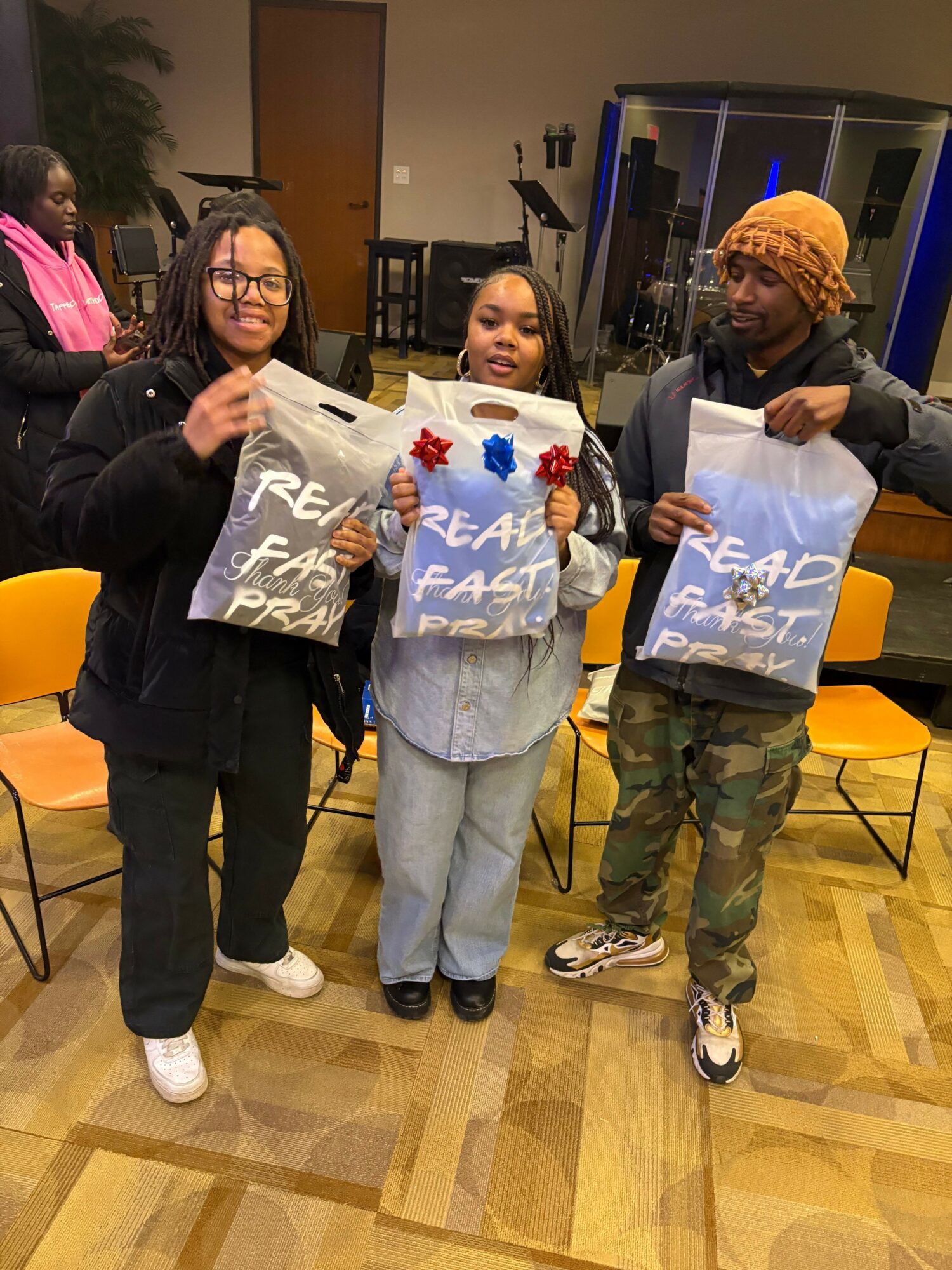 Three people holding signs, standing indoors on a wooden floor, with a person in the background wearing a beanie and camouflage pants.