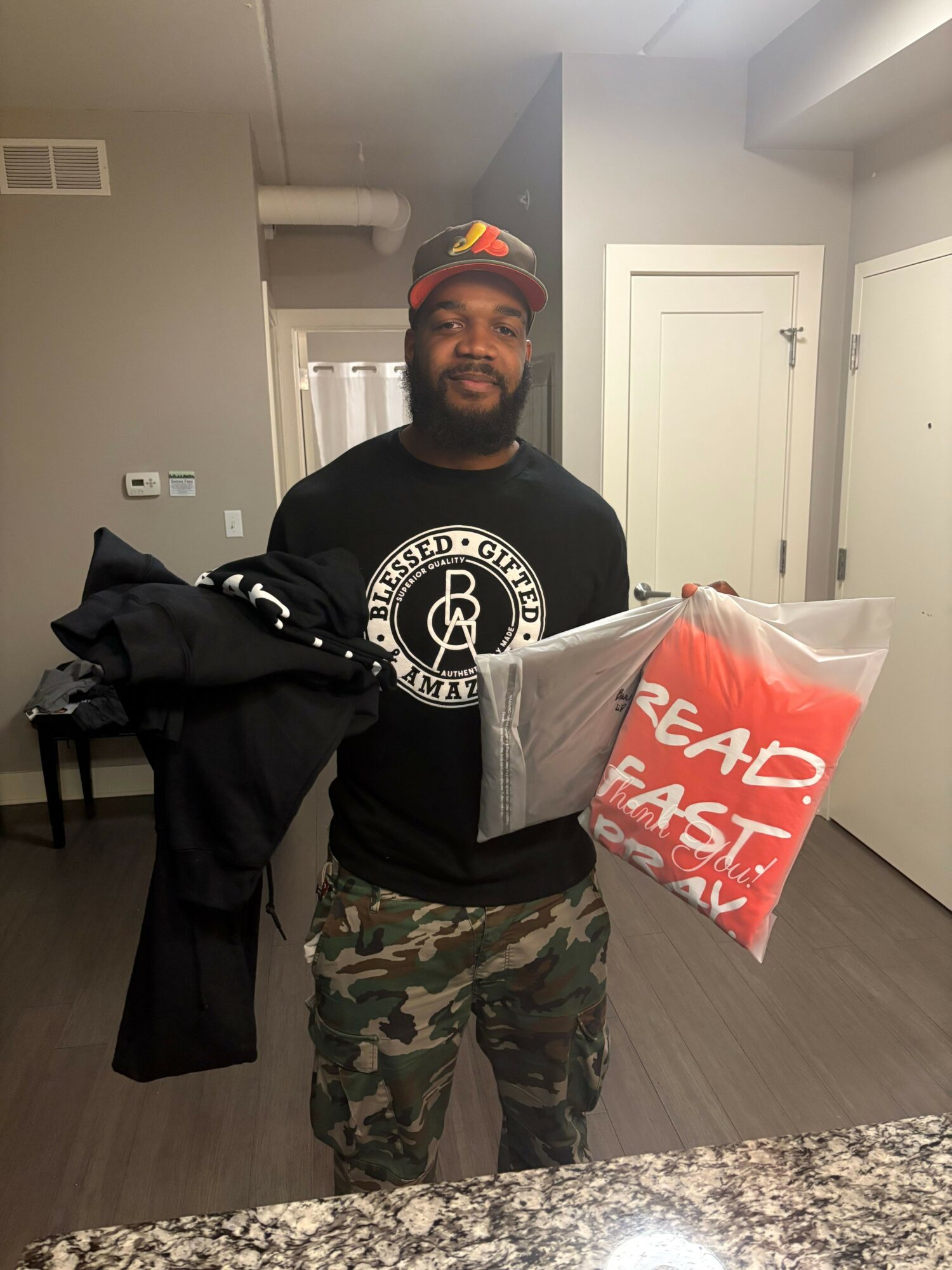 Man holding a large bag with red and white text, wearing a black shirt with a logo, camouflage pants, and a colorful cap.