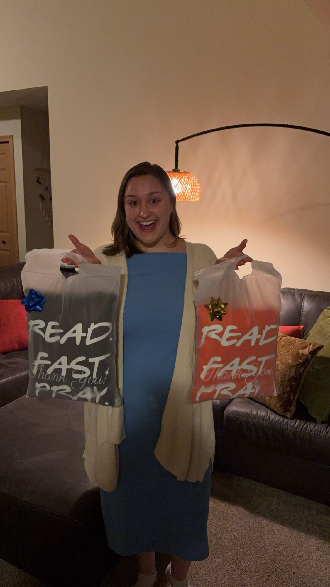 Person standing in living room holding two bags, smiling, with a lamp and sofa in background.