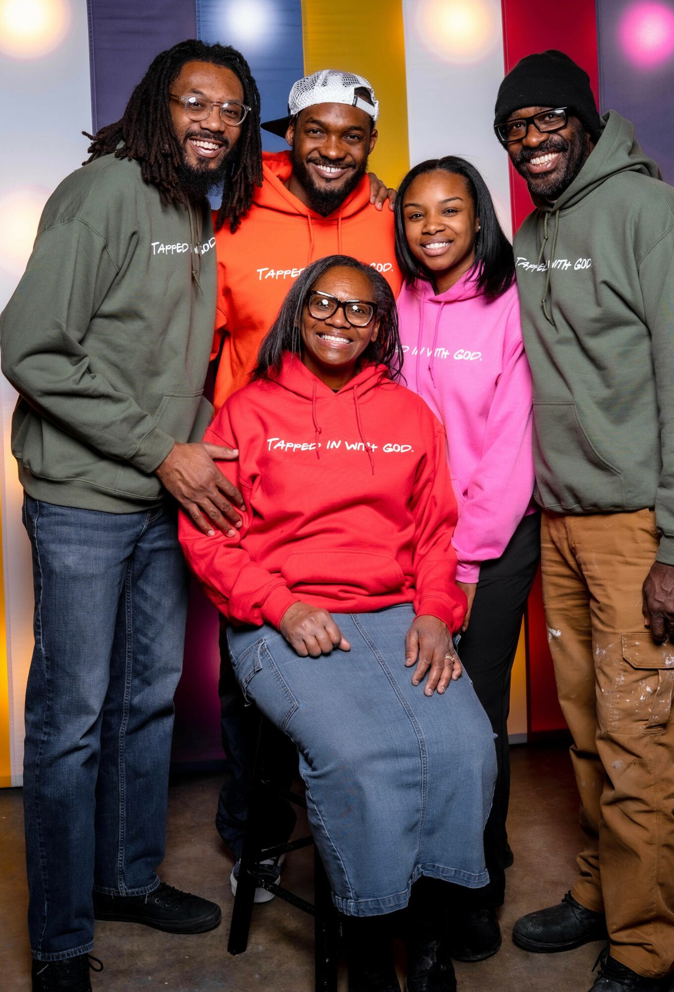 Group of five smiling people, three men and two women, standing together in colorful background, wearing casual hoodies and glasses.