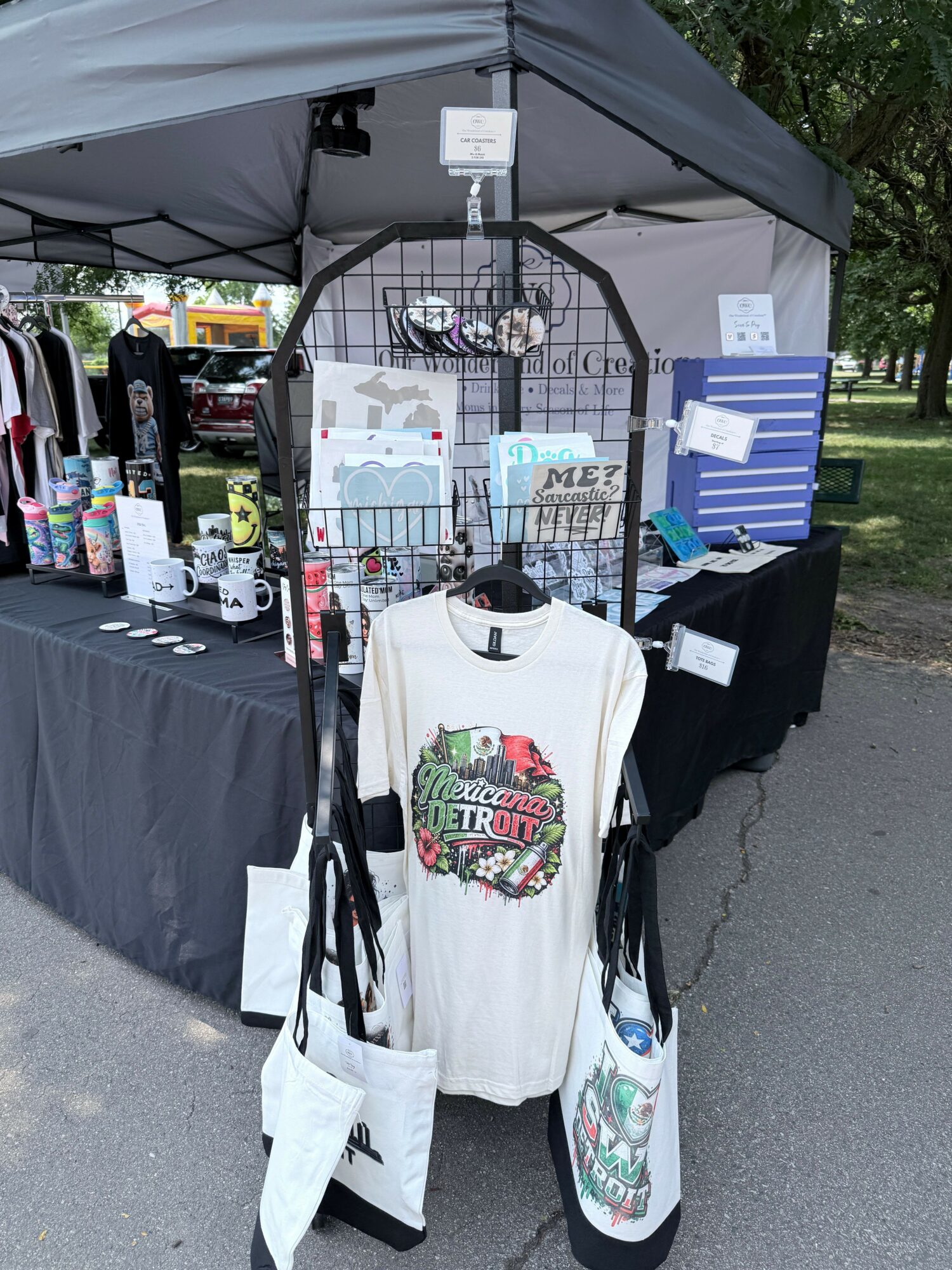 Display booth with clothing and accessories under a canopy, featuring a white T-shirt with a colorful graphic, and various items on tables.
