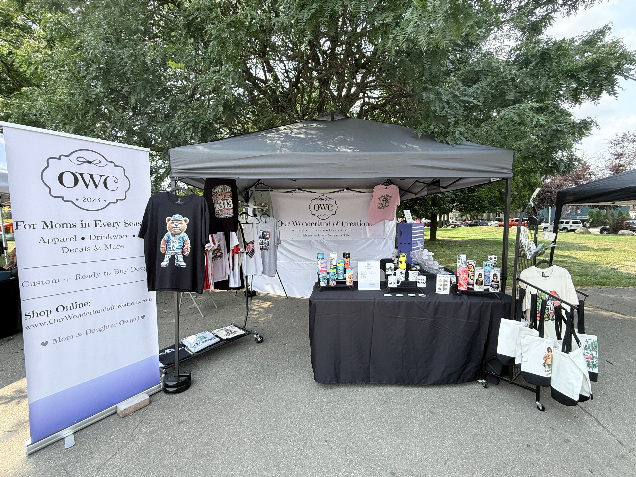 Outdoor market stall with a canopy, displaying clothing, skincare products, and informational materials, set in a park with trees and grass.