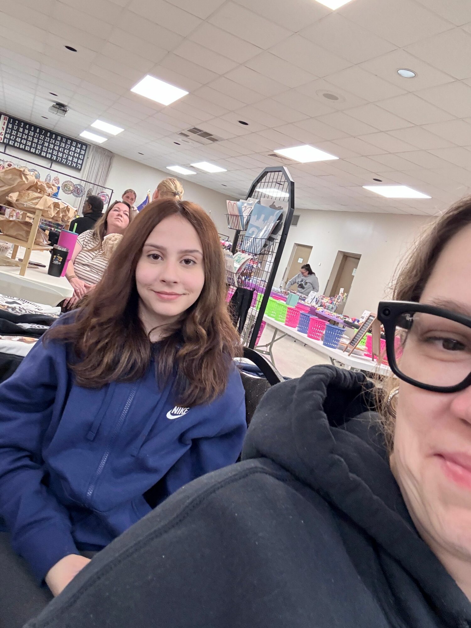 Two women sitting indoors, one wearing glasses and a dark hoodie, the other with long brown hair in a blue jacket, smiling.