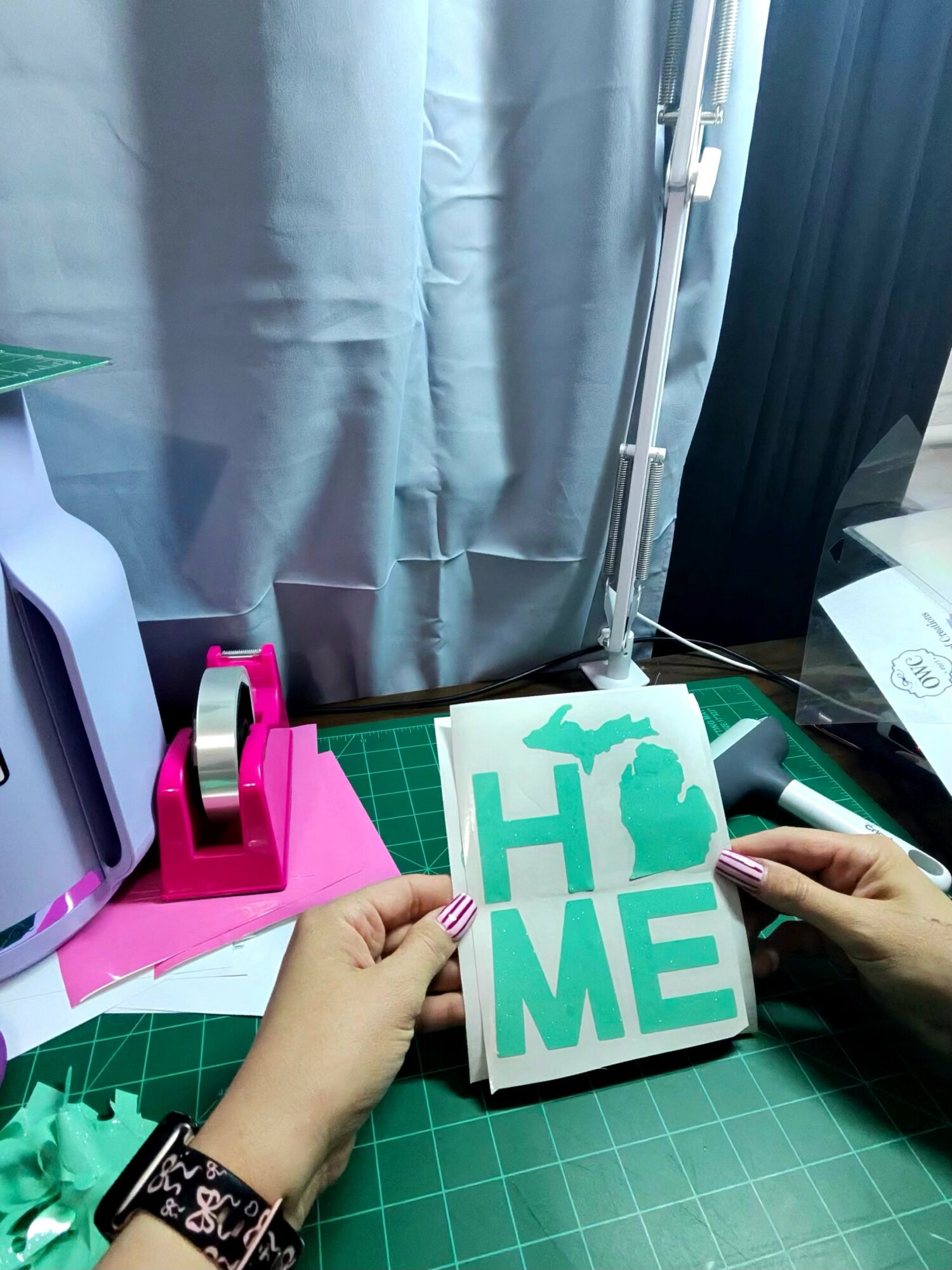Person holding a card with the word 'HOME' in teal letters, workspace with cutting mat, tape dispenser, and curtains in background.