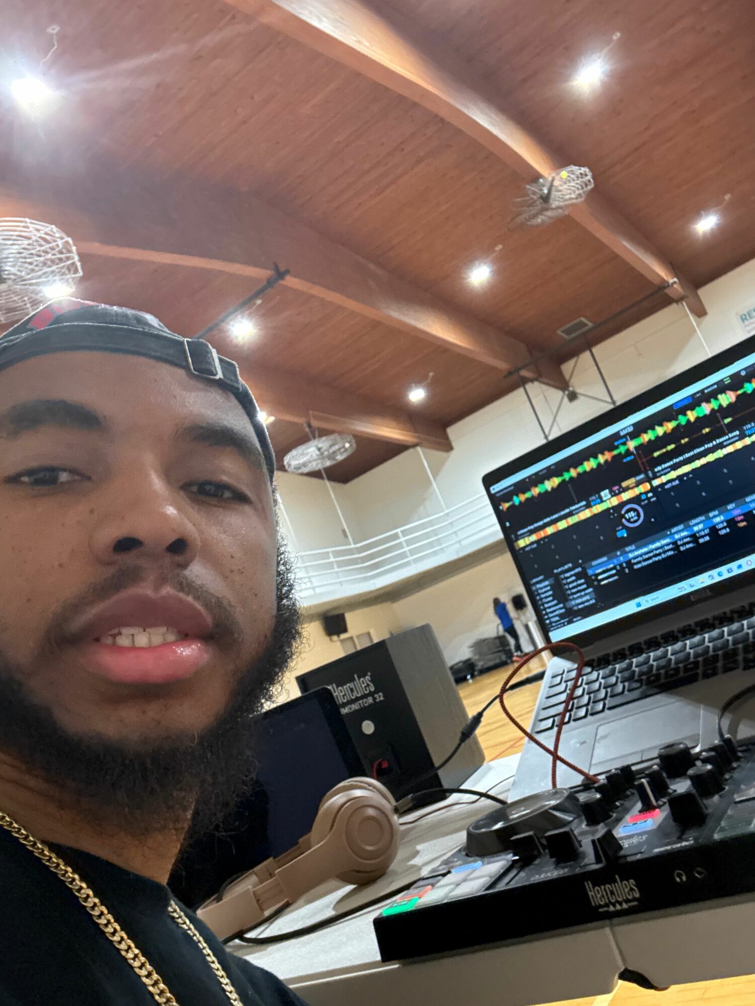 Person with a beard and cap smiling in a room with a computer and music equipment.