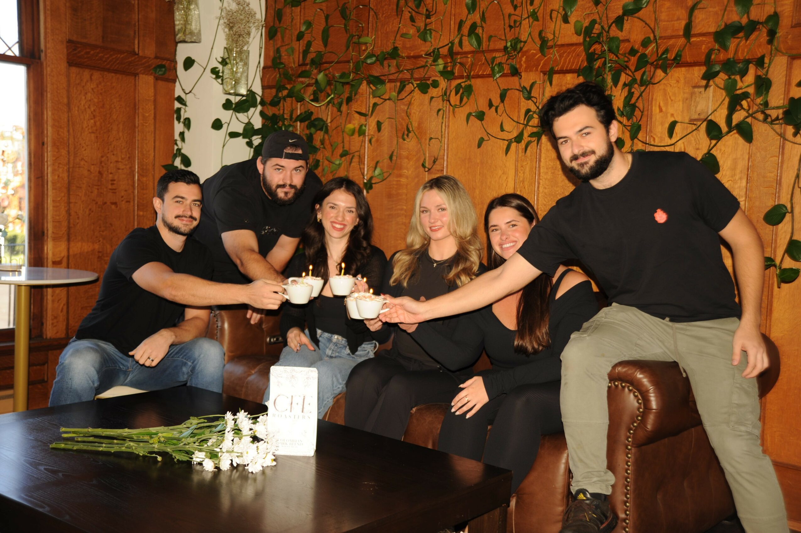 Group of six people sitting on a couch, raising glasses in a toast, in a cozy room with wooden walls and plants.